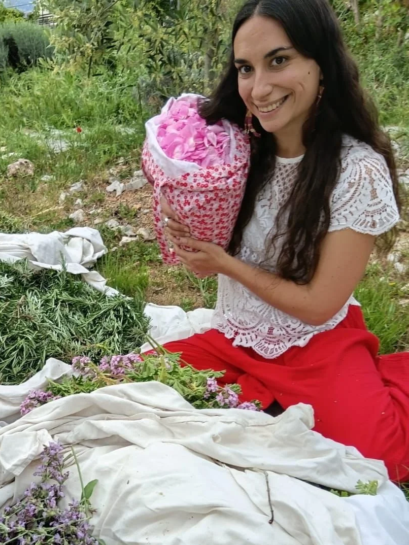 Une femme souriante assise dans un champ, tenant un grand sac de pétales de fleurs roses, entourée de végétation et de plantes vertes.