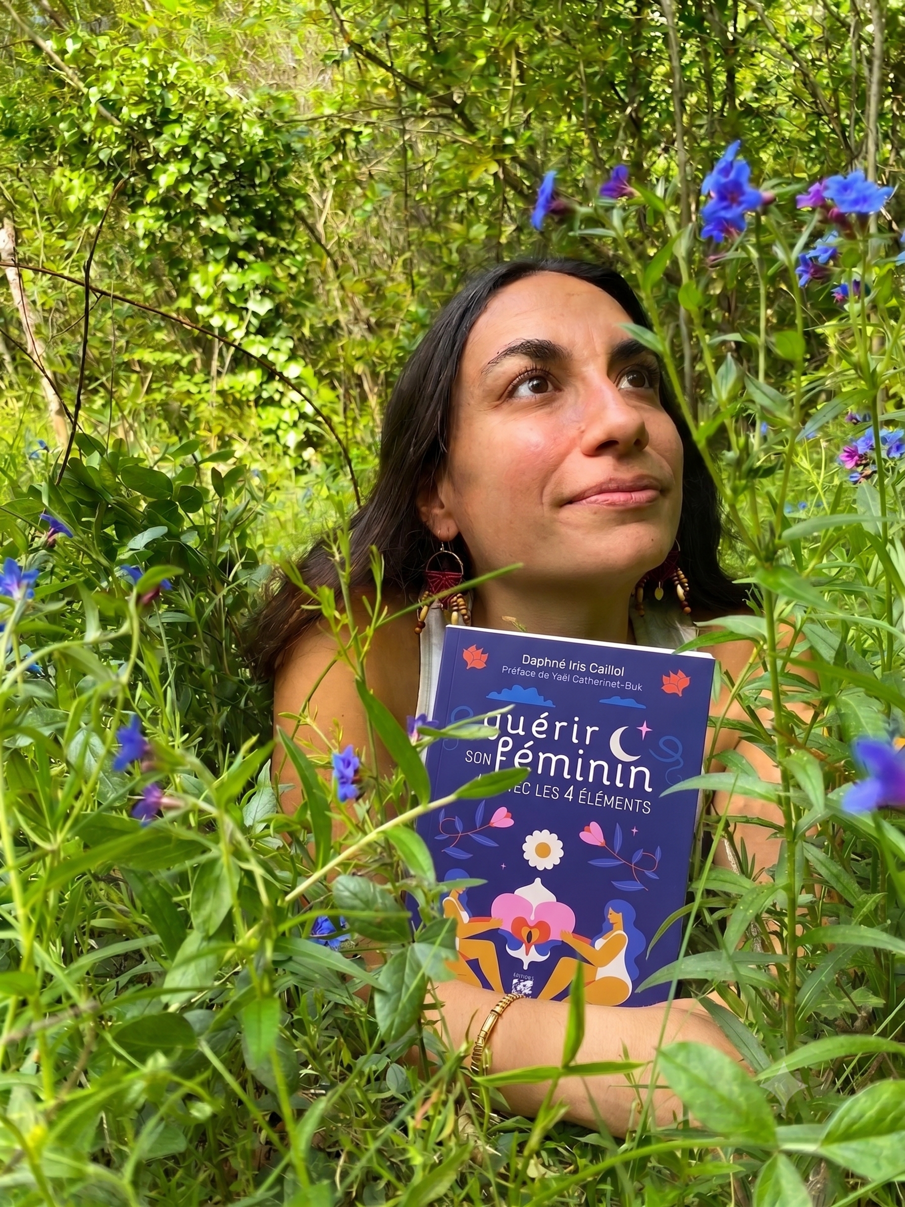 Femme avec des cheveux longs noirs, portant des boucles d'oreilles et un bracelet, tenant un livre bleu s'intitulant : "Guérir son féminin avec les 4 éléments" de Daphné Iris Caillol