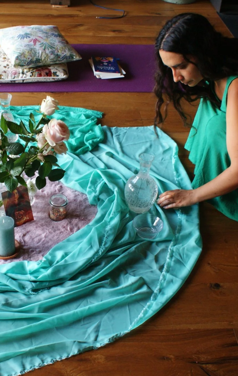 Une femme ajuste une nappe turquoise sur un sol en bois, avec des fleurs roses en vase, des bougies et des objets de décoration, dans un espace intérieur. préparation de la retraite pour femmes