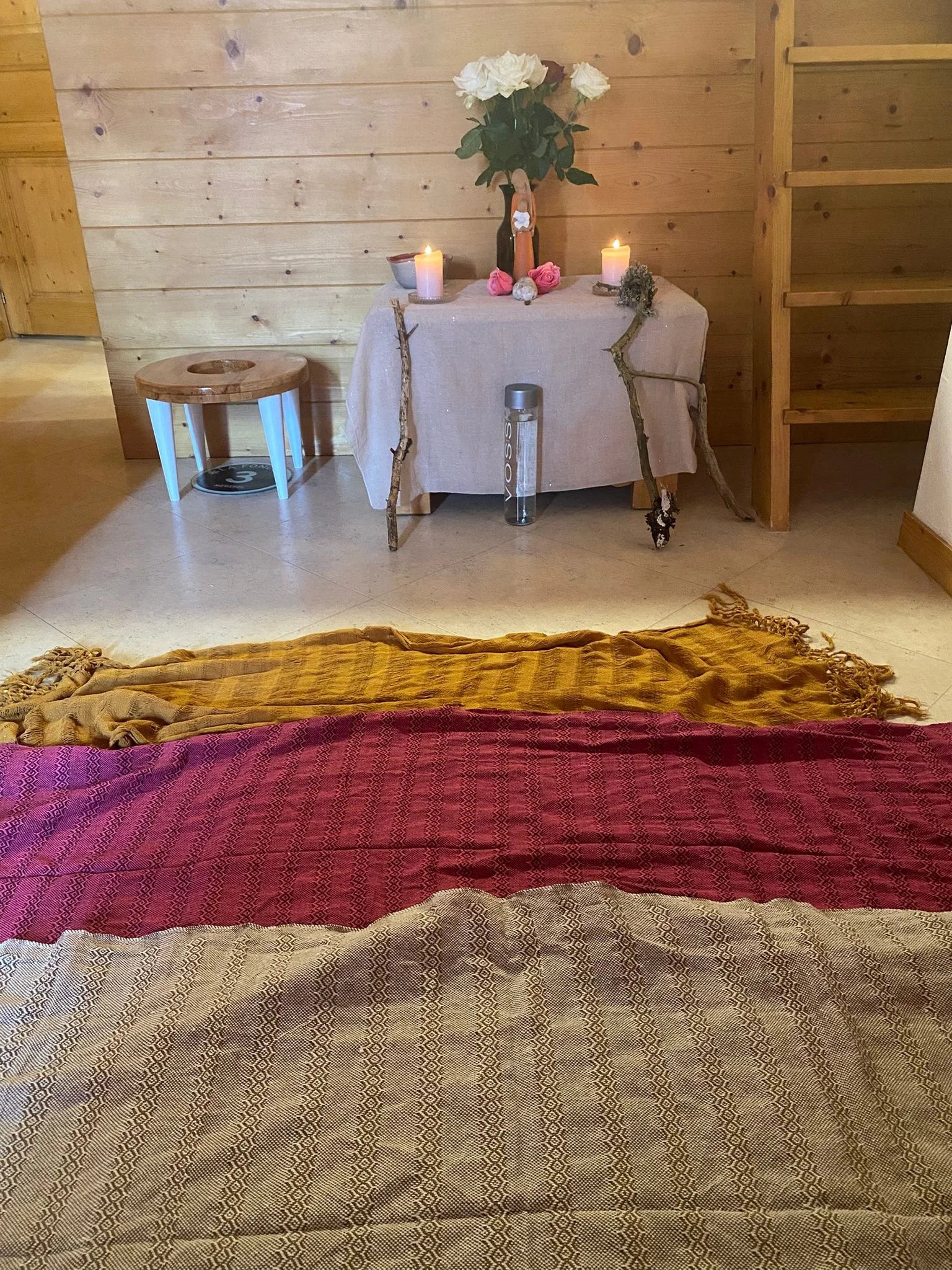 Region intérieure avec un lit fait de tissus à motifs, dans une pièce en bois, vue partielle sur une table avec des fleurs, bougies et branches, et une bouteille d'eau.