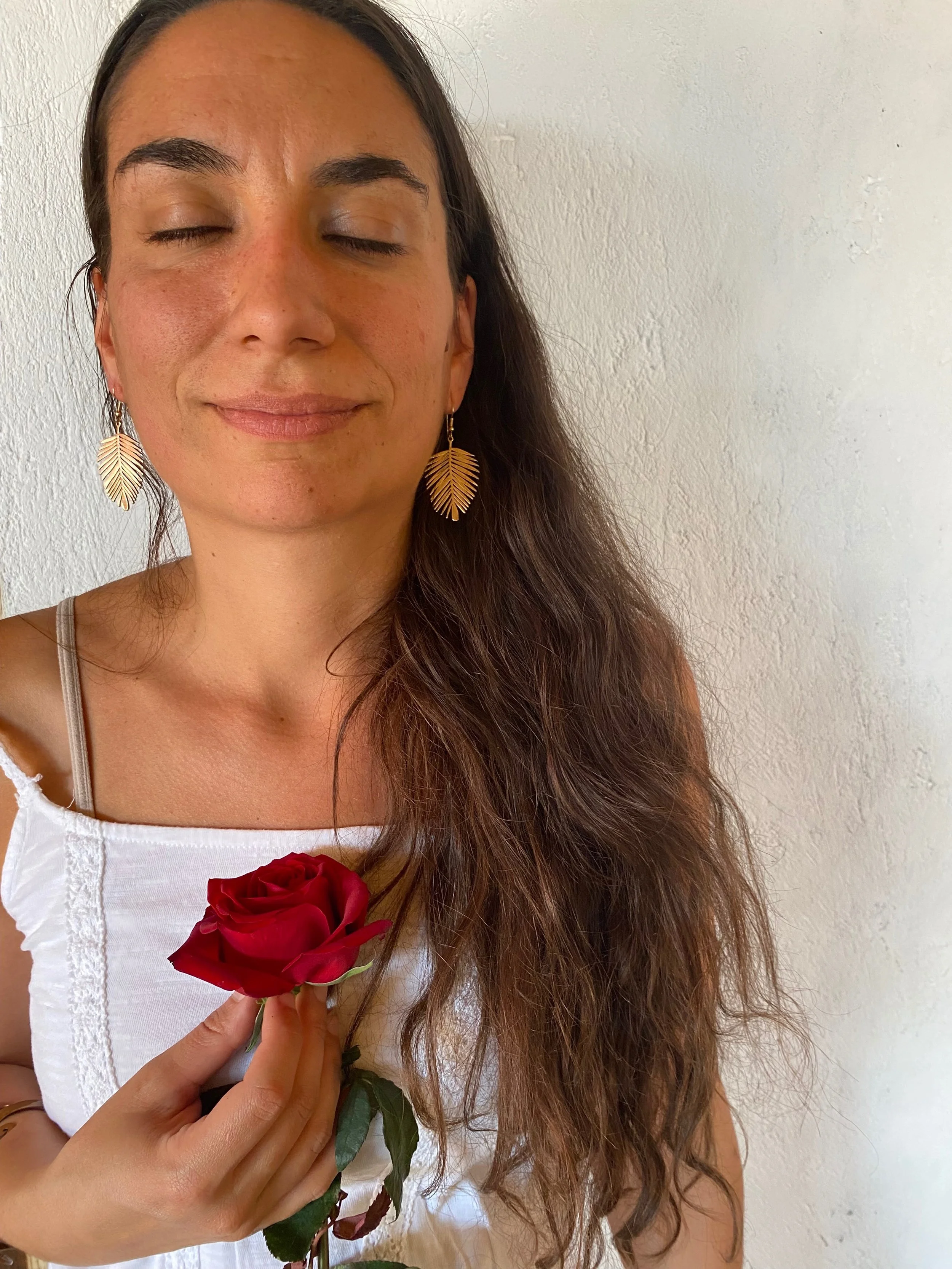 Une femme avec de longs cheveux bruns, portant des boucles d'oreilles en forme de feuilles, tenant une rose rouge.