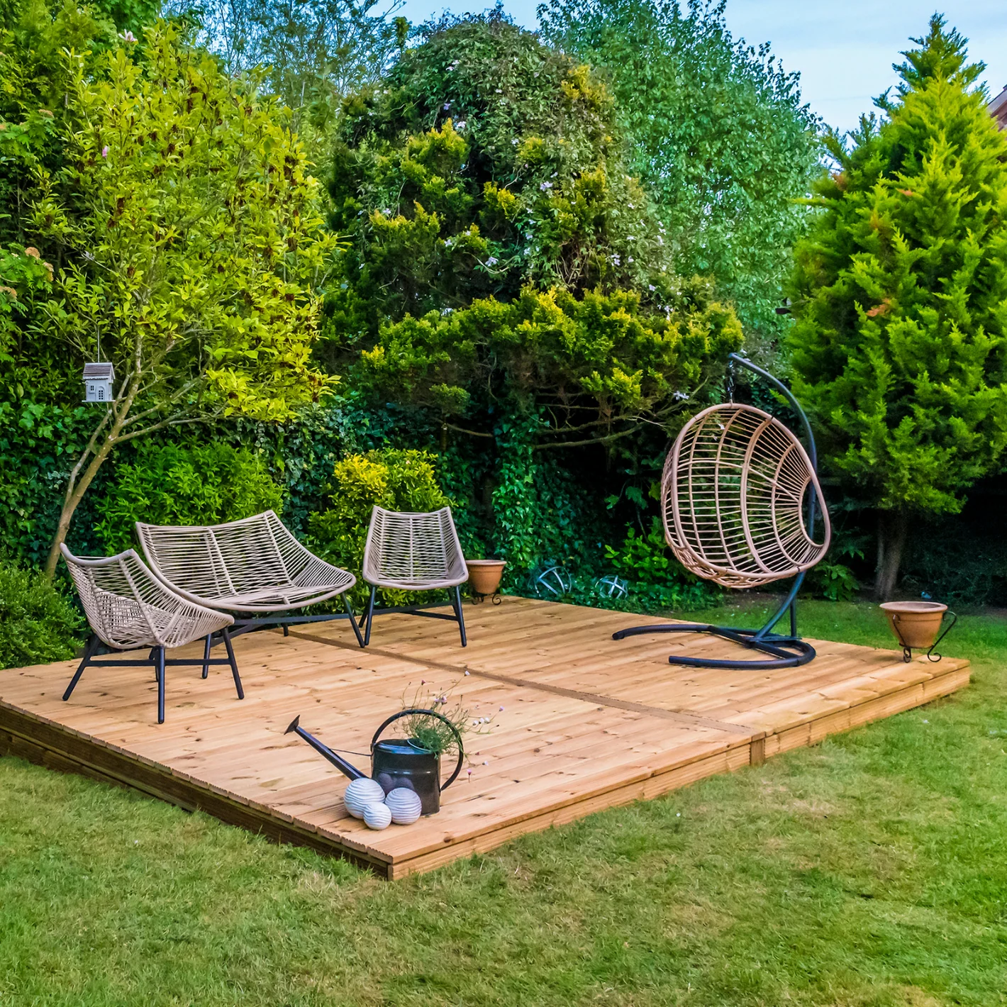 A backyard garden with a wooden deck, outdoor chairs, a hanging egg chair, and gardening tools surrounded by lush green bushes and trees.