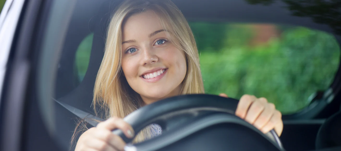 A young woman with blonde hair smiling as she sits behind the steering wheel of a car, looking out the windshield.