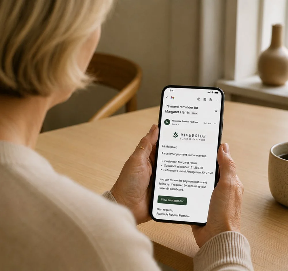 A woman holding a smartphone showing an email from Riverside Funeral Partners about an overdue payment for Margaret Harris.