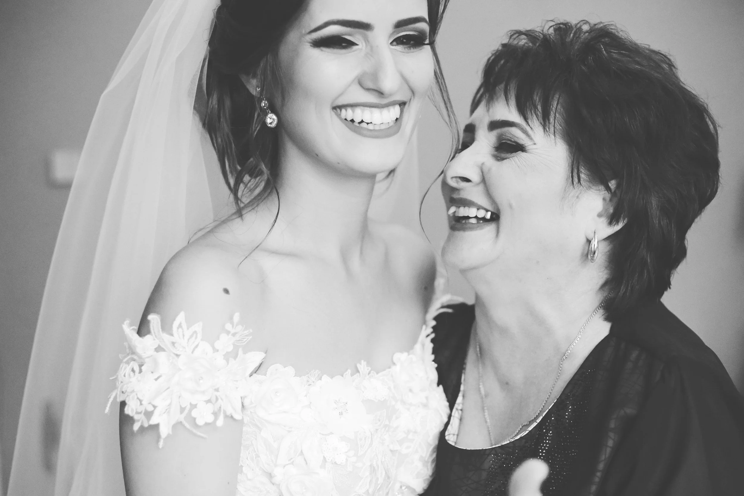 A black and white photo of a bride in a wedding dress smiling with a woman, possibly her mother, who is also smiling and looking at her.