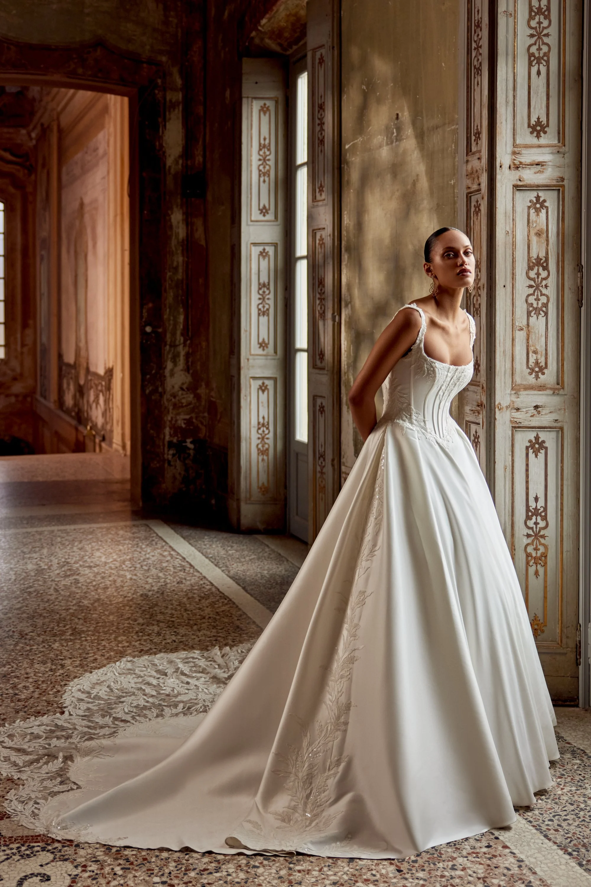 A woman in a white wedding gown standing inside a historic building with tall windows and ornate wooden panels on the walls.