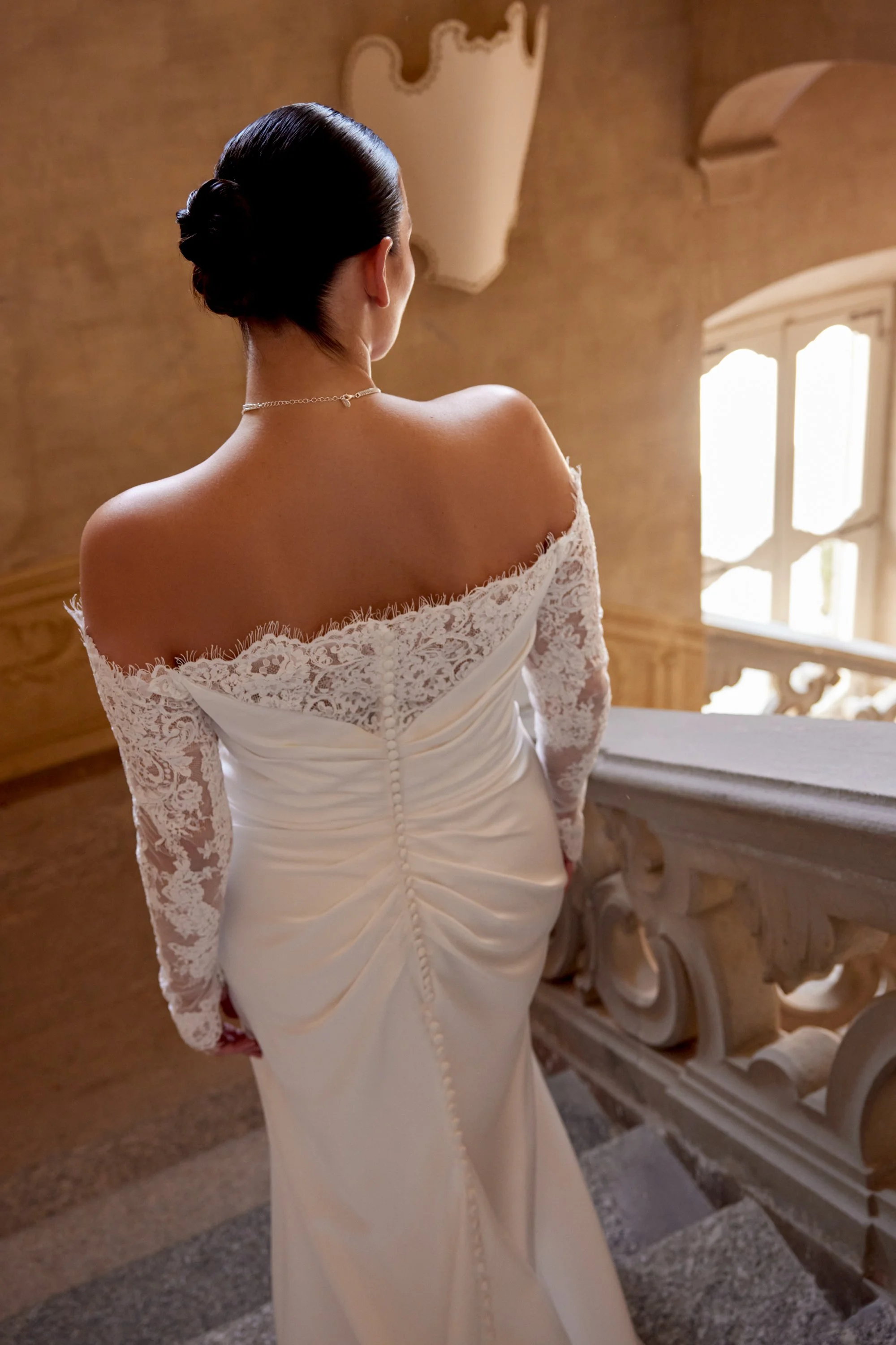 A woman dressed in a lace and satin wedding gown with her hair styled in an elegant updo, standing indoors near a staircase and a window, seen from behind.