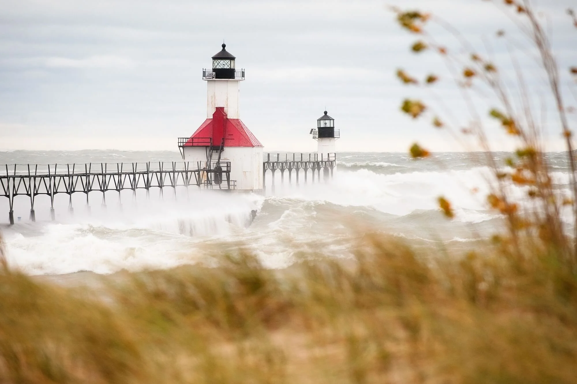 Waves crash against a white lighthouse sits at the end of a pier.