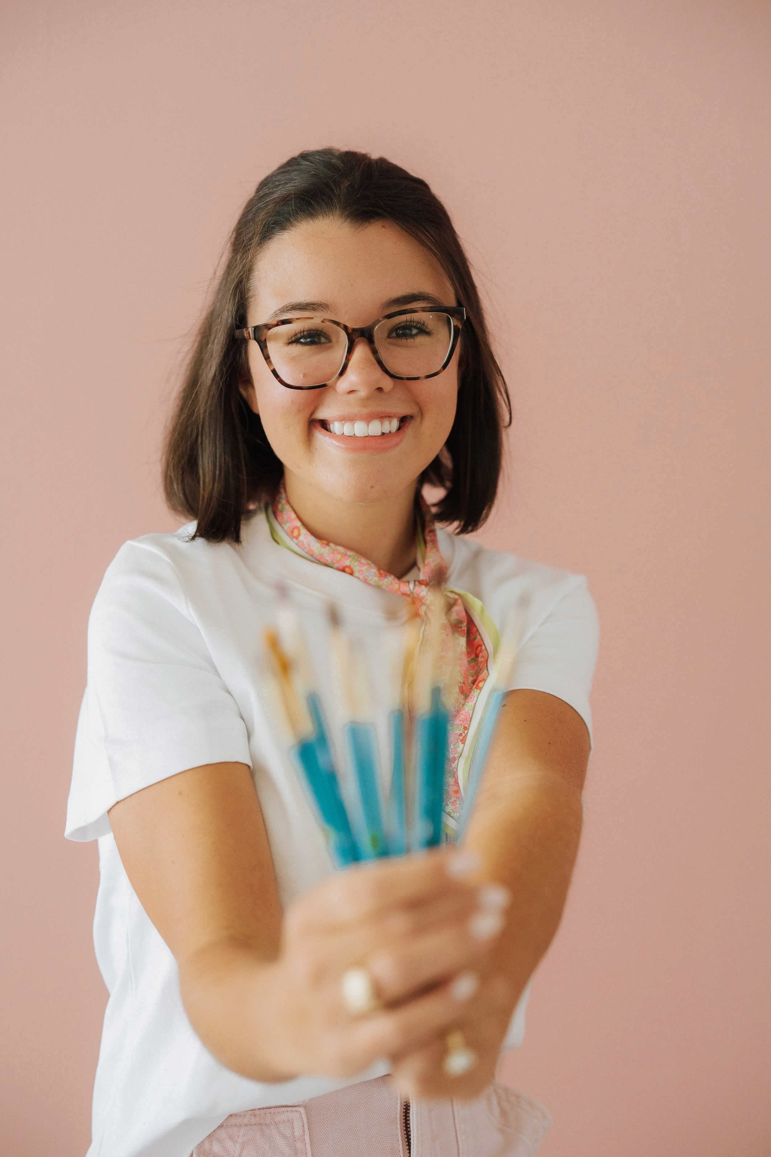 Young woman with brown hair, glasses, and a bright smile, holding in her hand a colorful bundle of pens towards the camera against a pink background.