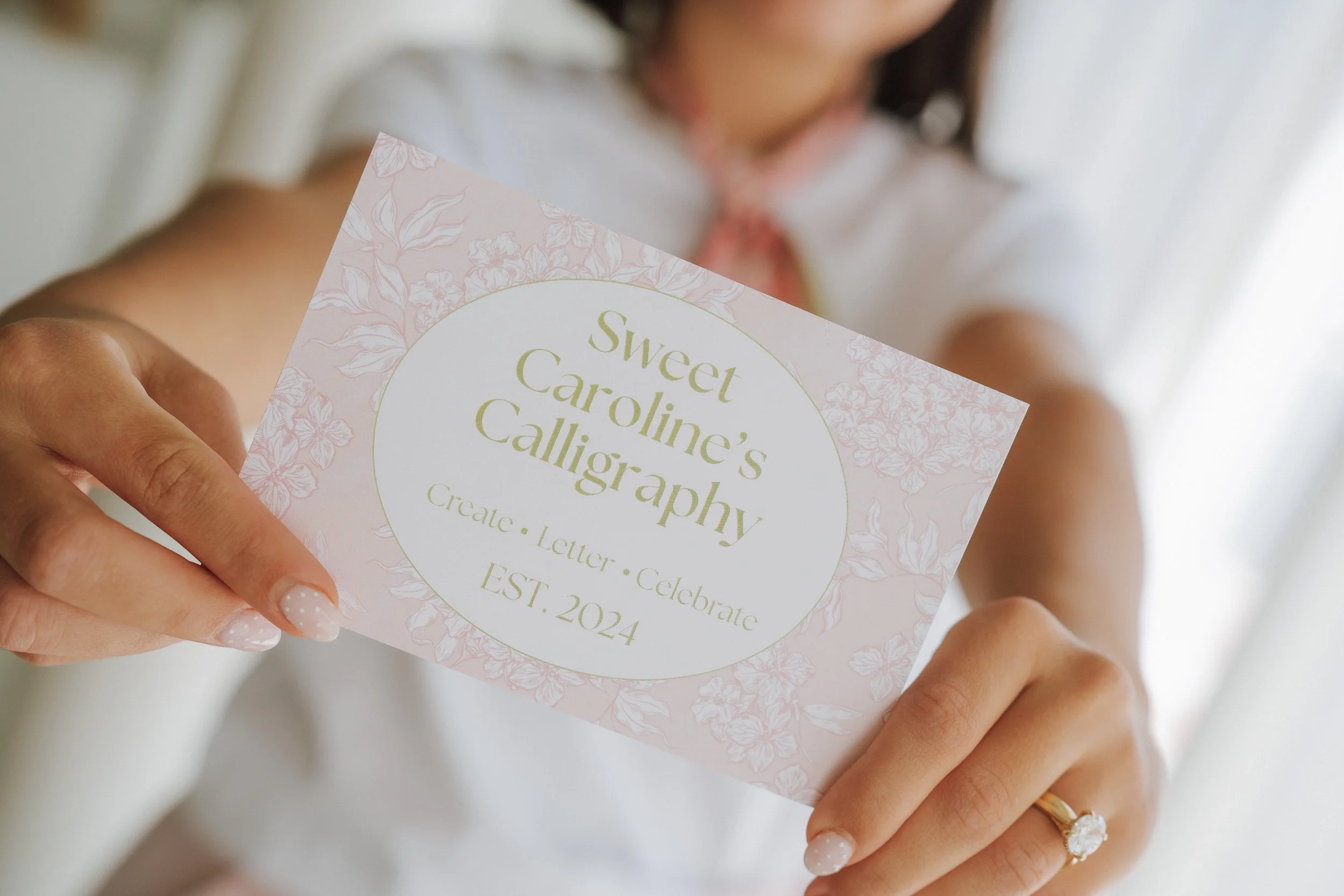 A woman holding a pink floral-patterned card with the text "Sweet Caroline's Calligraphy" and details about creating, lettering, and celebrating, with the date established in 2024.