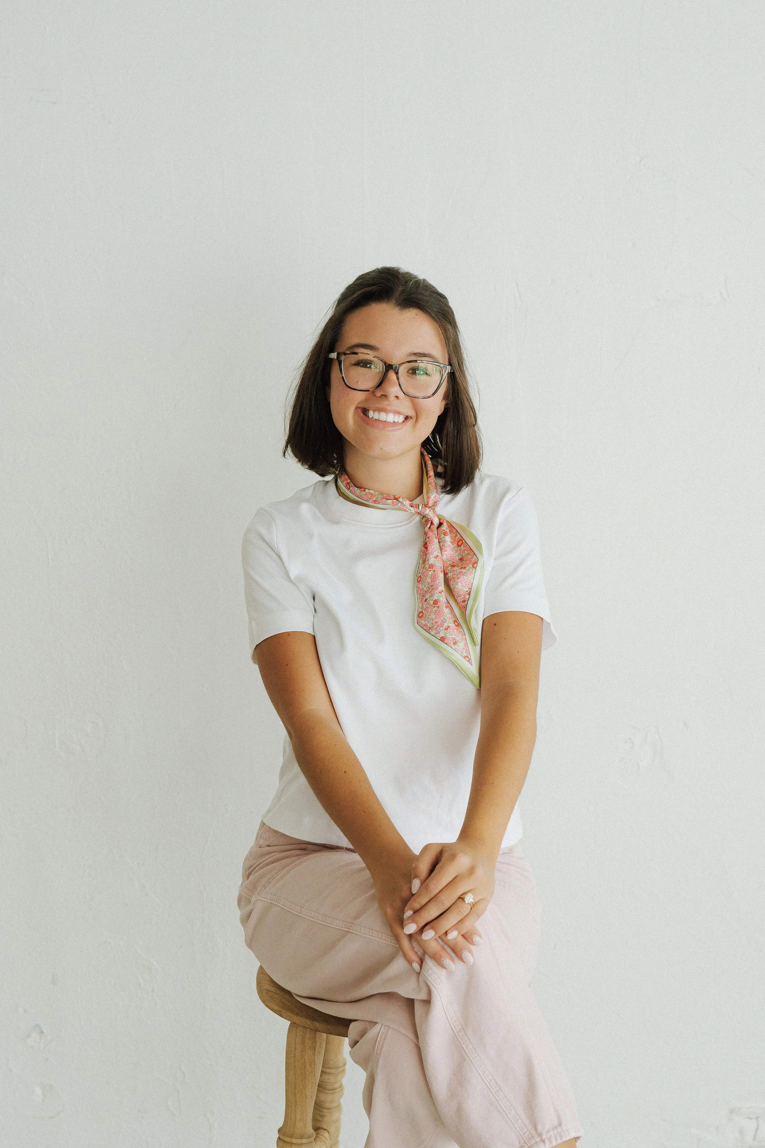 A young woman with shoulder-length dark hair, glasses, and a cheerful smile, sitting on a wooden stool against a plain white wall. She wears a white top, a pink and green patterned scarf, and beige pants.