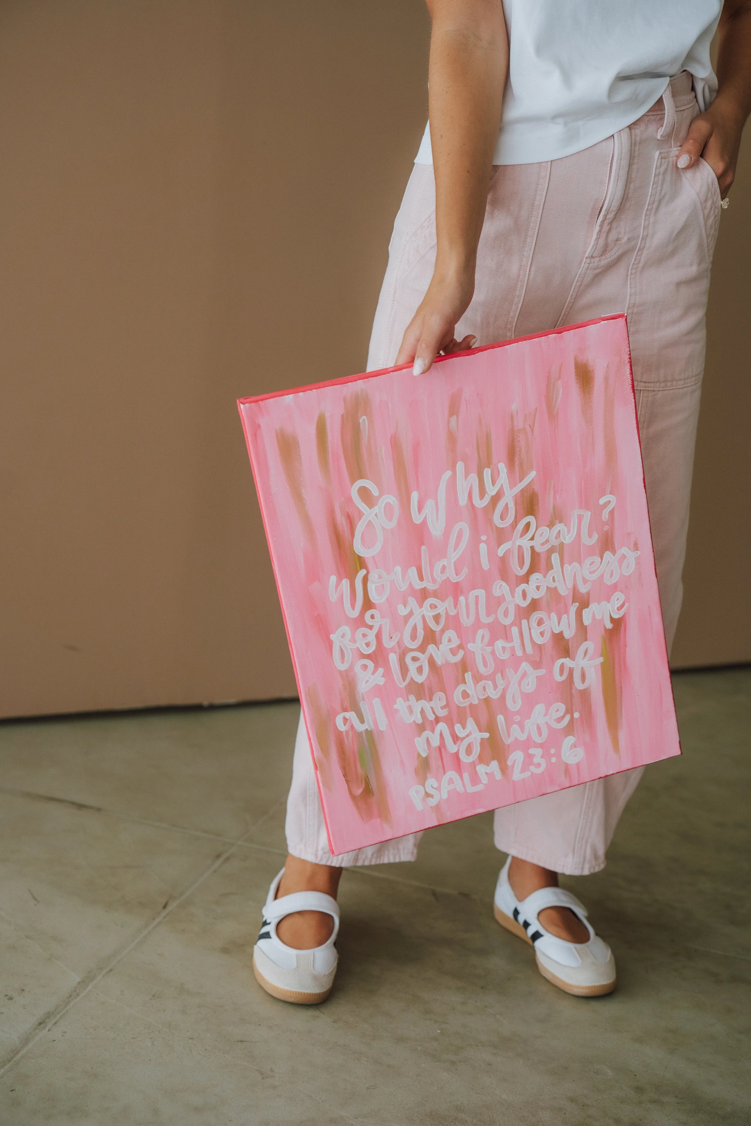 A person wearing a white shirt and light pink pants, standing on a tiled floor, holding a pink sign with white handwritten scripture text.
