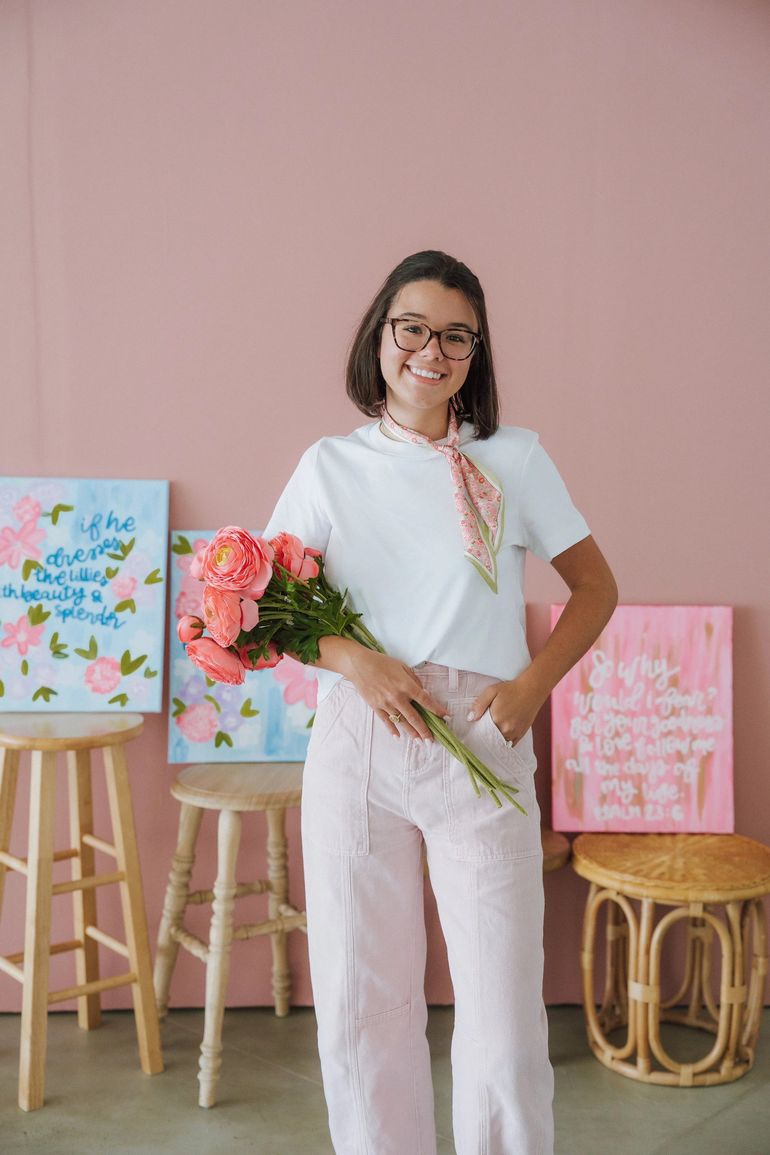 A woman with glasses smiling while holding a bouquet of pink flowers inside a pink-colored room with art posters and wooden furniture.