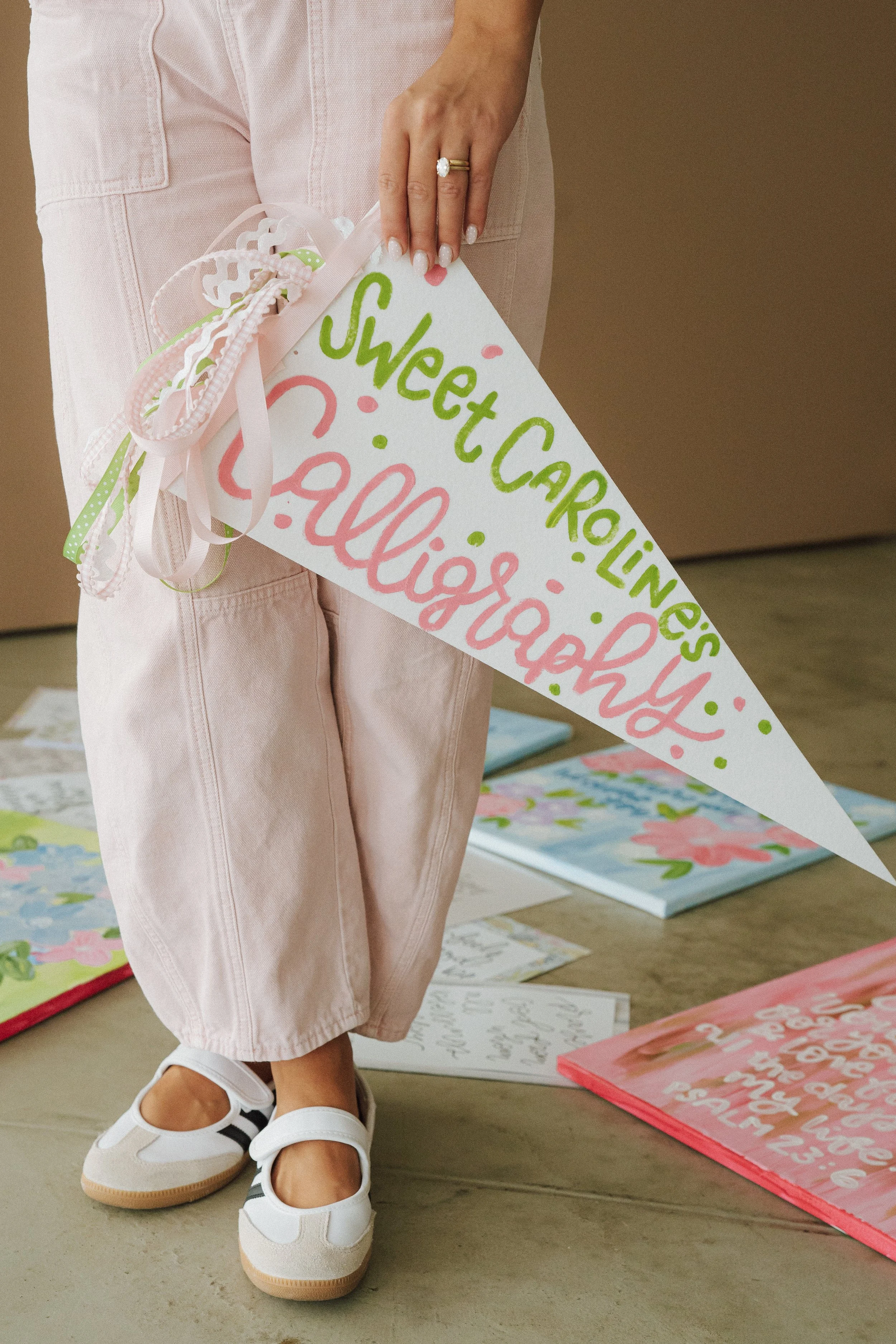 A person holding a white triangular sign with pink and green handwritten text with pink and green decorative dots, standing on a wooden floor surrounded by colorful, floral-themed posters or artwork laid flat.