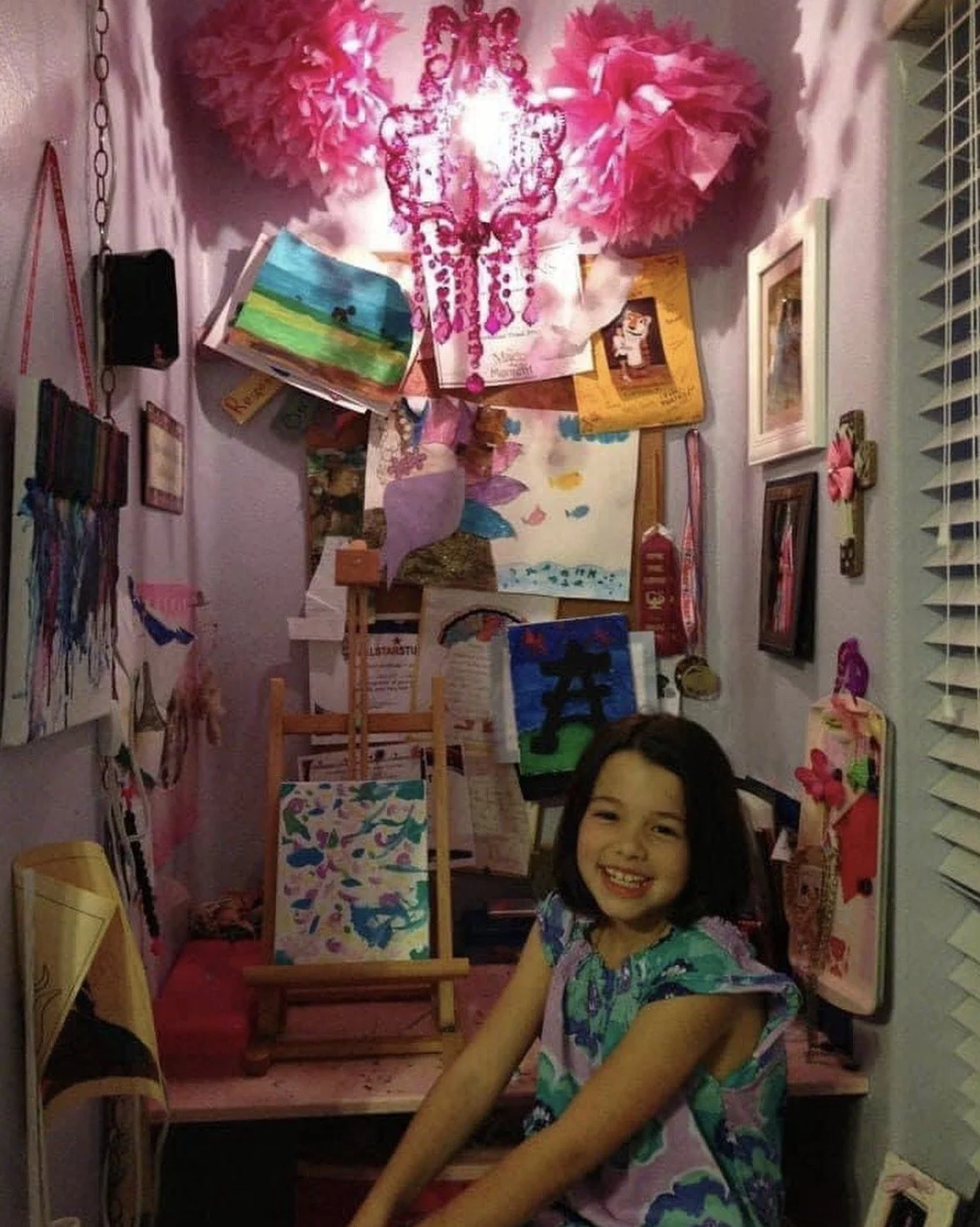 A young girl smiling in a colorful art studio surrounded by paintings, drawings, and art supplies with pink paper pom-poms and decorations on the wall.