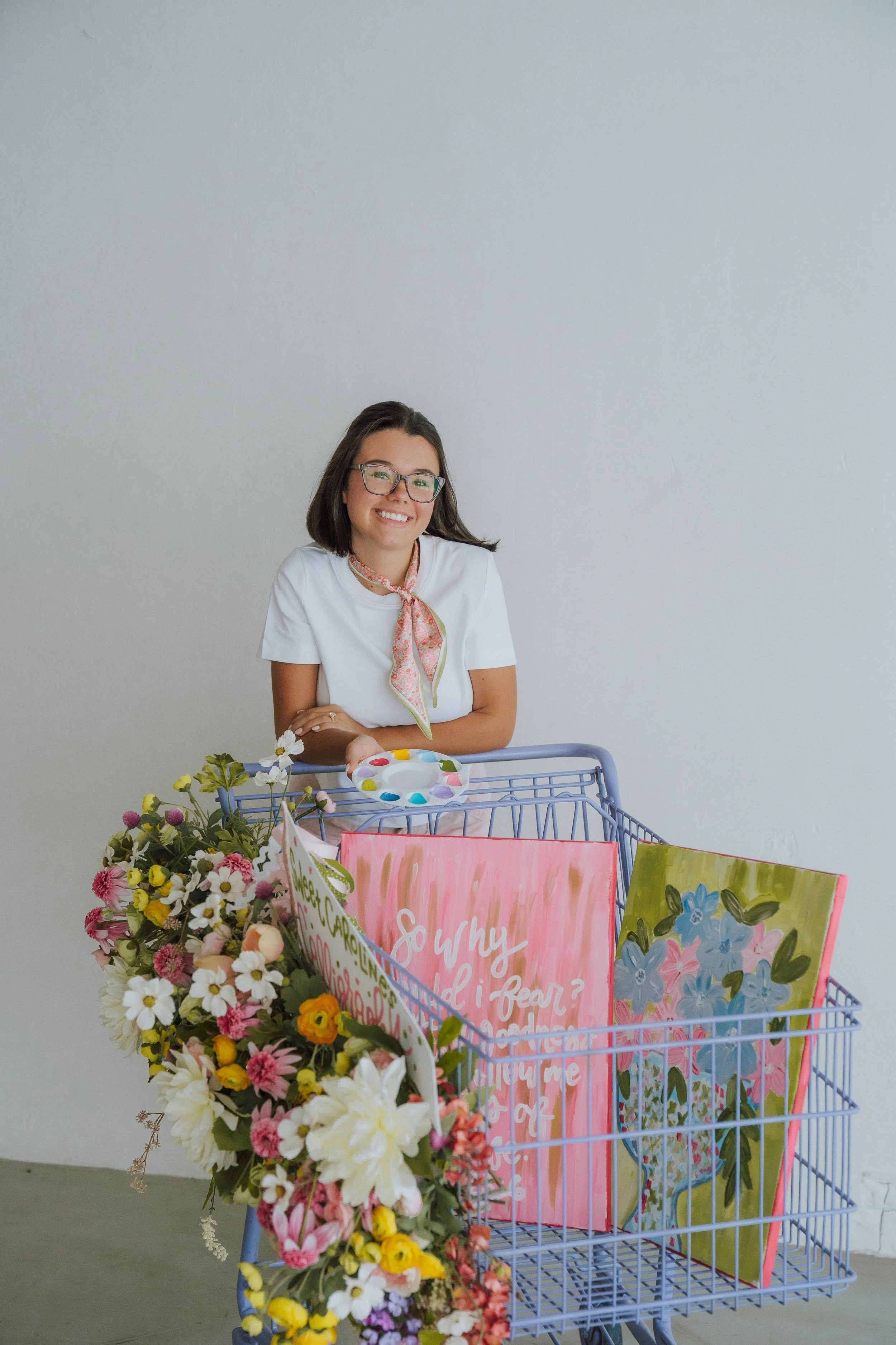 A smiling woman with glasses and dark hair, wearing a white shirt and a colorful scarf, is sitting behind a shopping cart filled with colorful flowers and banners with inspirational messages.