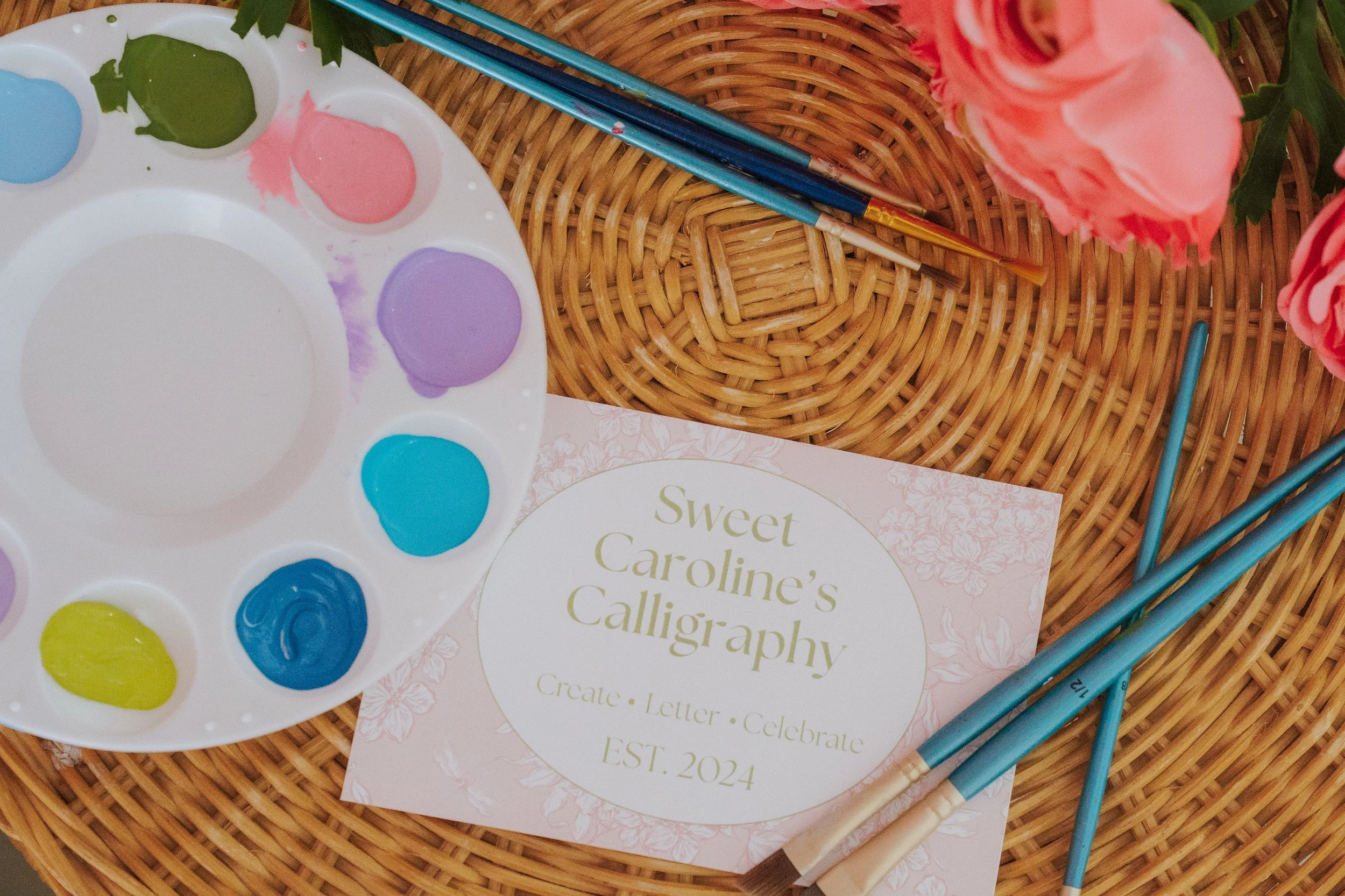 A watercolor paint palette with blue, purple, green, pink, yellow, and other colors, placed on a wicker surface. Next to it are watercolor brushes, a pink flower, and a card that reads 'Sweet Caroline's Calligraphy'.