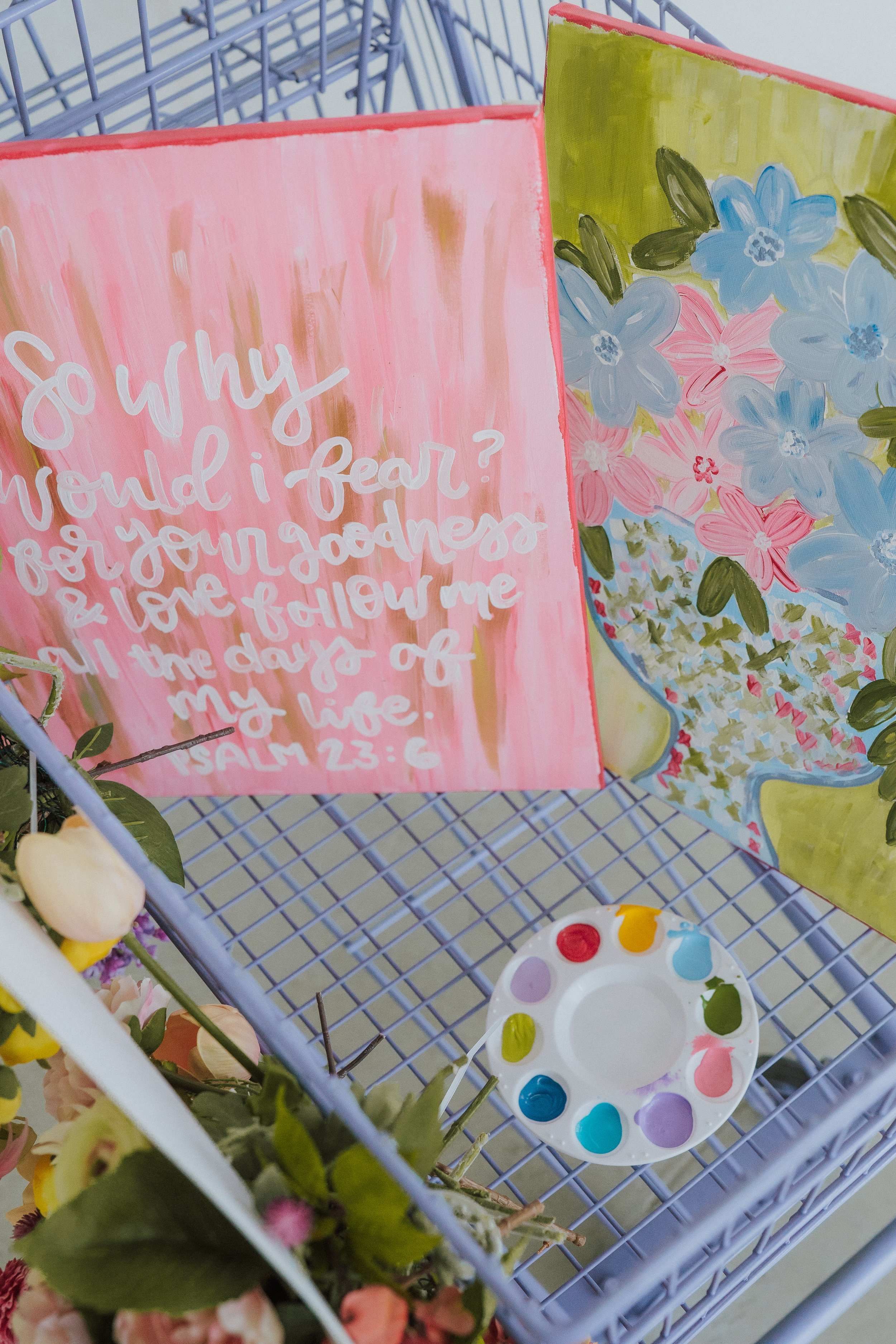 A painted pink card with a heartfelt message resting on a metal shopping cart, surrounded by floral arrangements including pink and blue flowers. A white ceramic palette with colorful paints is also visible.