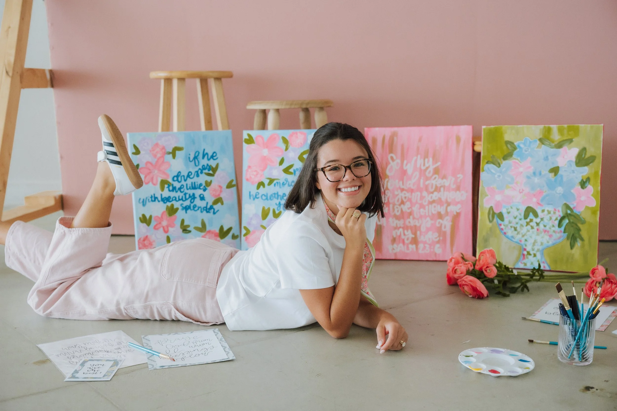 Young woman lying on the floor with a smile, gesturing towards her chin, surrounded by colorful paintings, art supplies, and pink roses in a bright studio with a pink wall and wooden stools in the background.