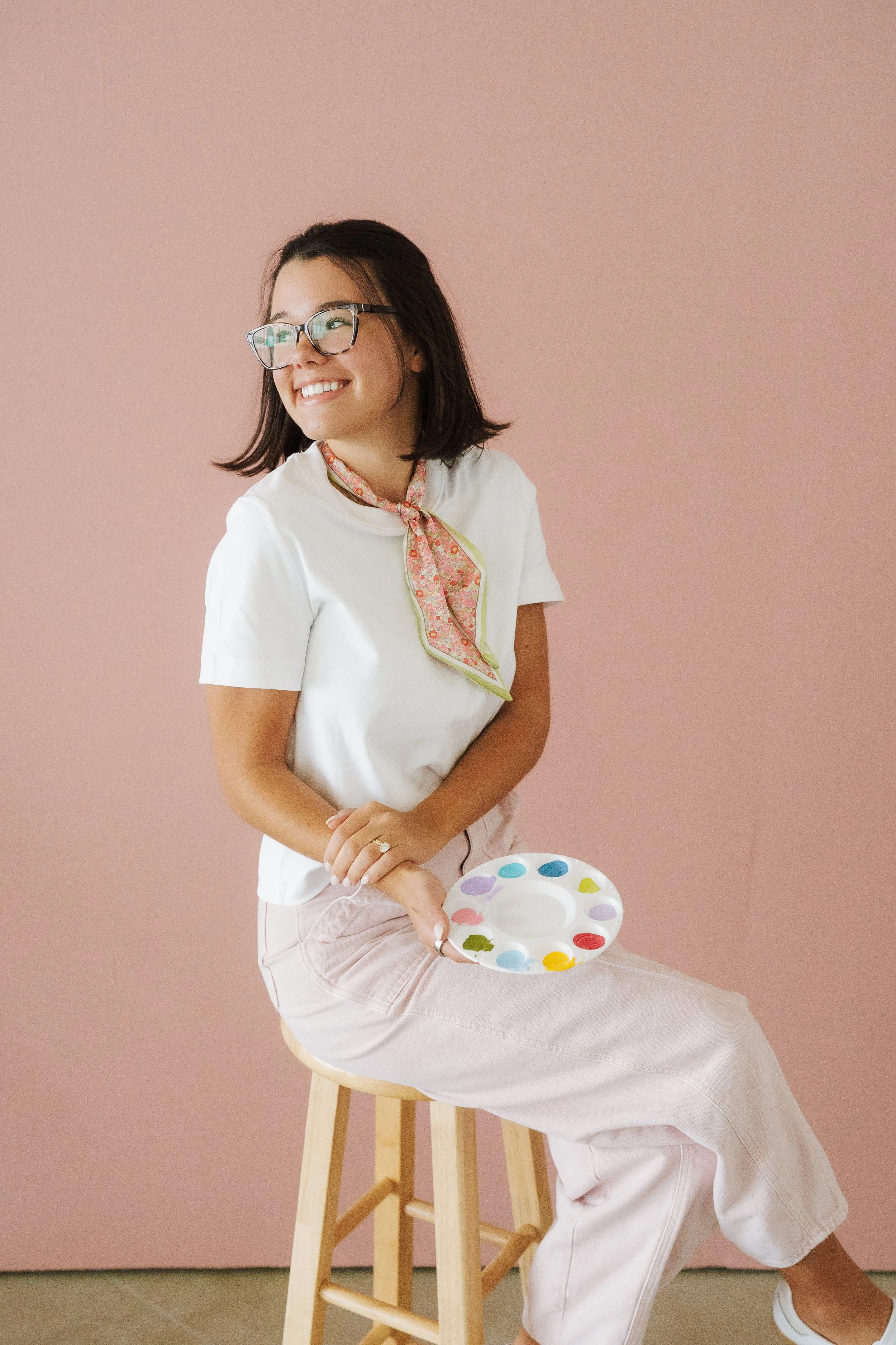 A young woman with shoulder-length dark hair, glasses, and a floral scarf around her neck, sitting on a stool against a pink wall, holding a paint palette.