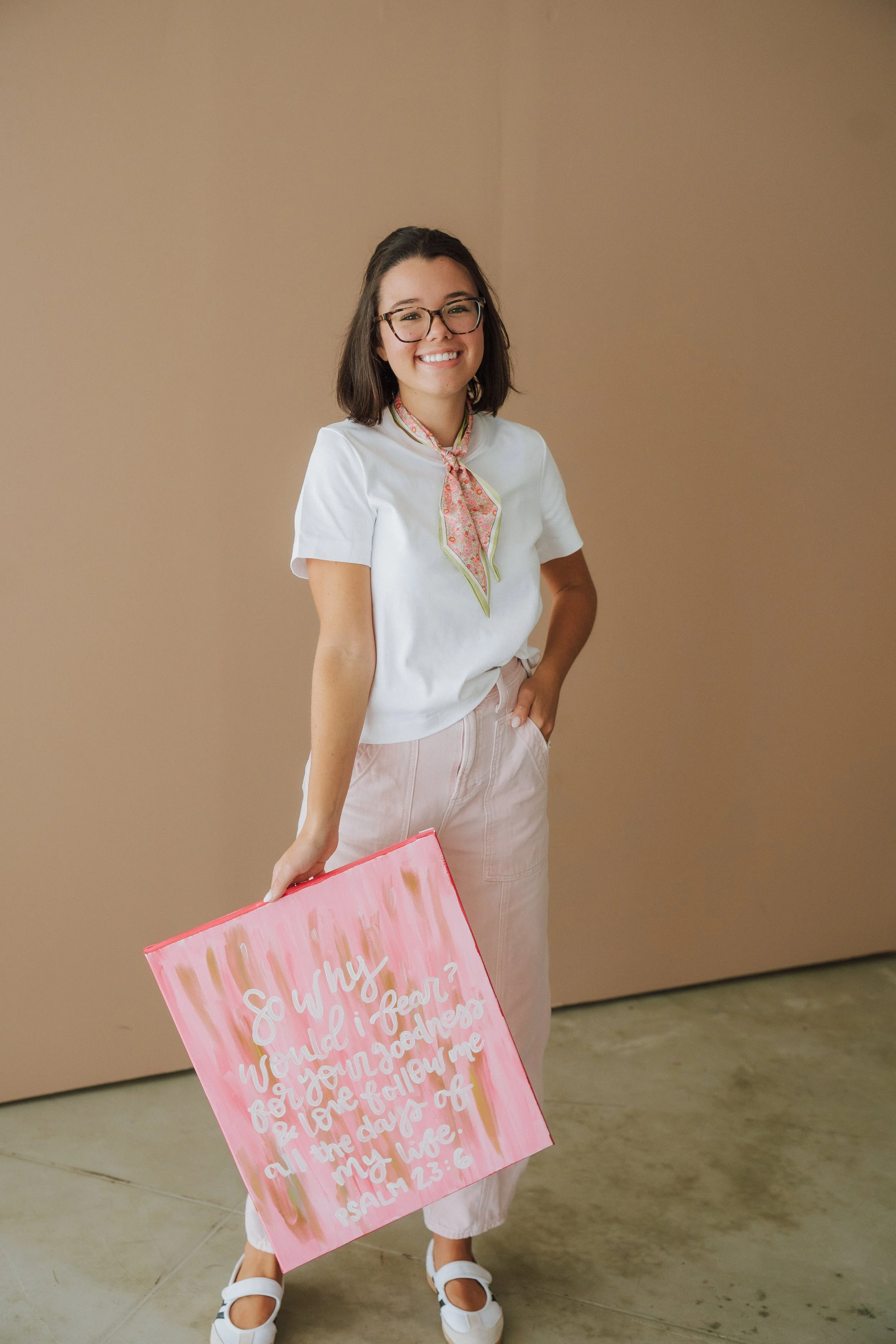 A woman with glasses, wearing a white shirt and pink skirt, holding a pink sign with handwritten text, smiling, standing in front of a plain beige wall.