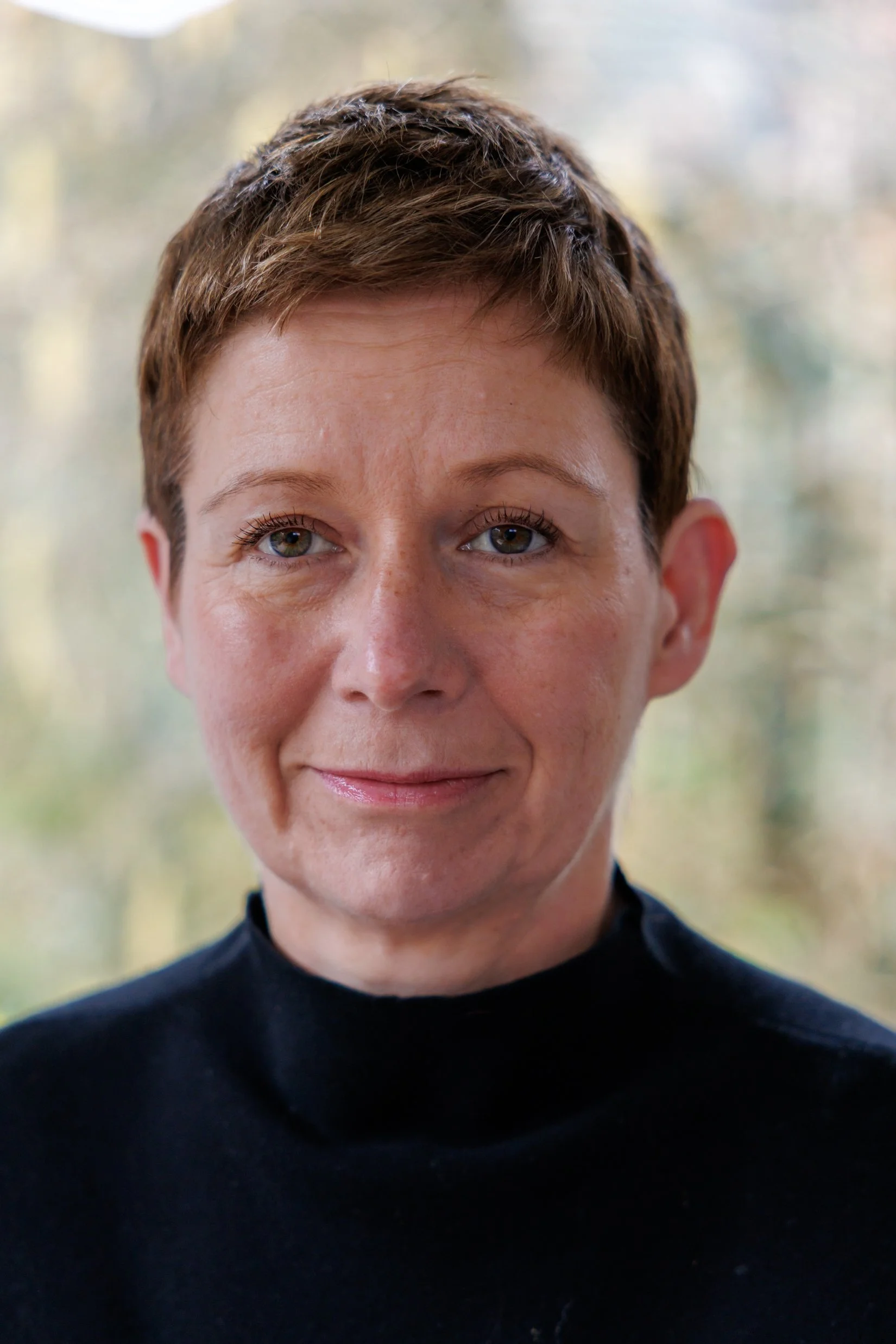 Close-up of a woman with short brown hair wearing a black top, smiling softly, with a blurred natural background.