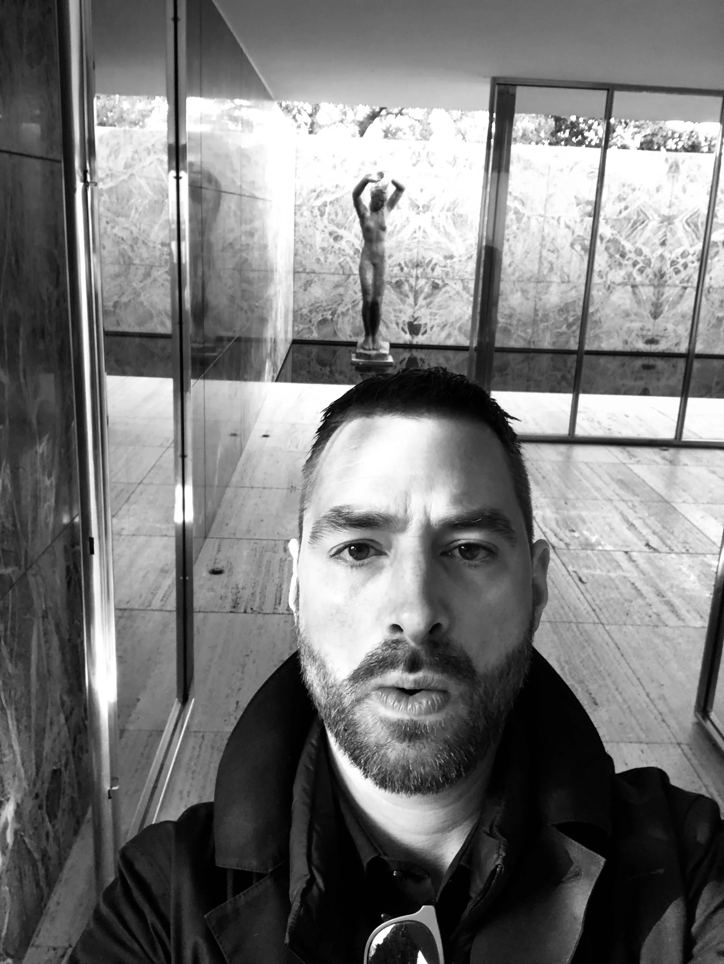 Black and white selfie of a man with a beard taking a photo in front of a mirror, with a sculpture of a woman with raised arms in the background.