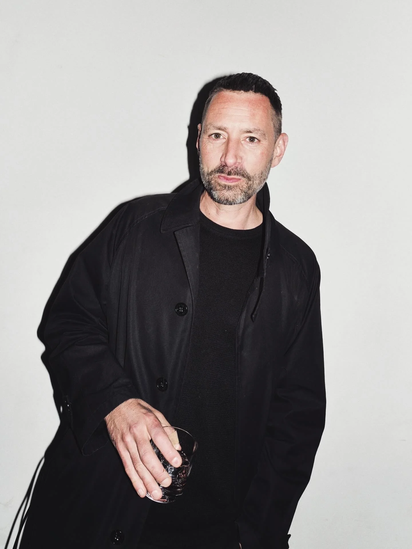 A man with a beard and short black hair, wearing a black coat and shirt, is holding a glass of dark liquid and standing against a plain white background.
