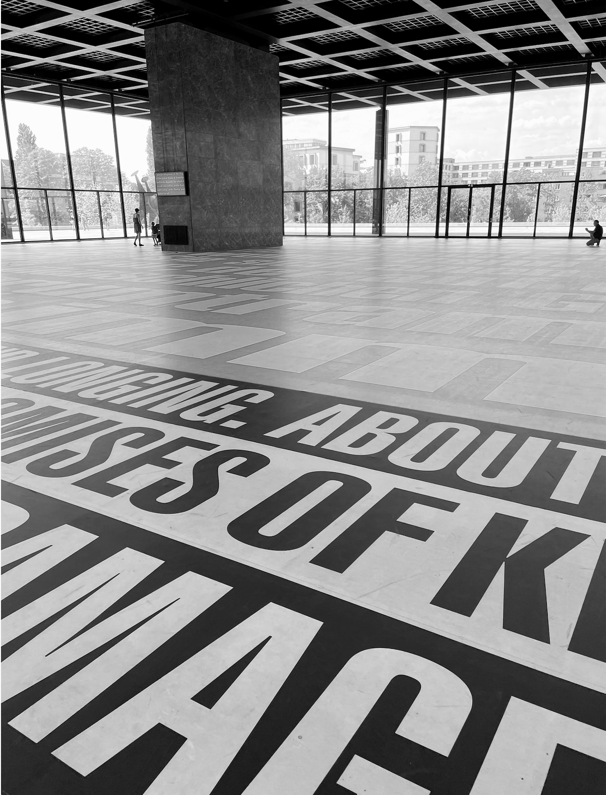 View of a spacious modern interior space with large floor-to-ceiling windows, a large rectangular column, and a ceiling grid with panels. There is text on the floor about requesting assistance.