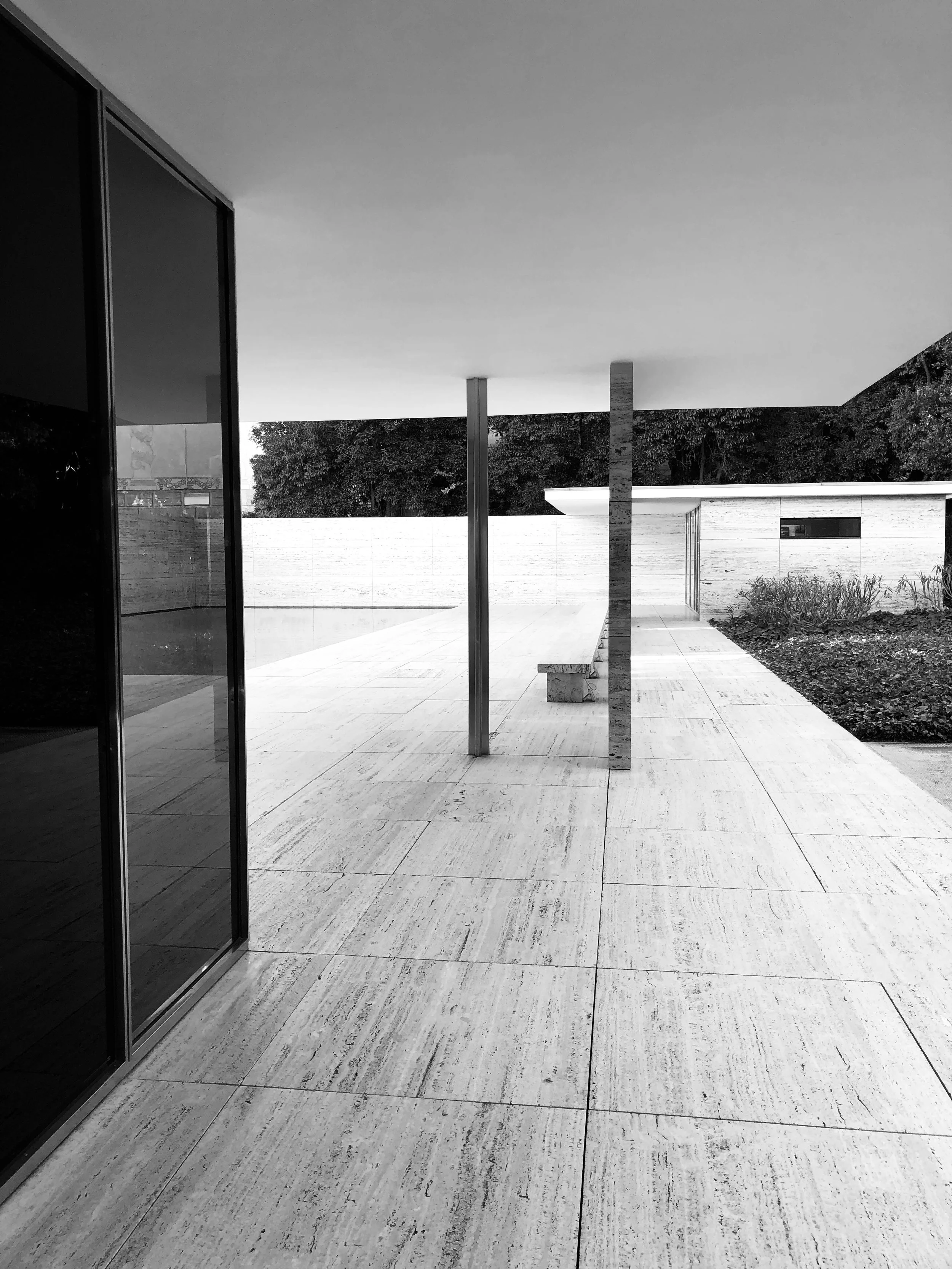 Modern outdoor patio with large tiled flooring, a glass wall on the left, metal and stone columns, and a minimalist building with garden to the right, under a flat roof.
