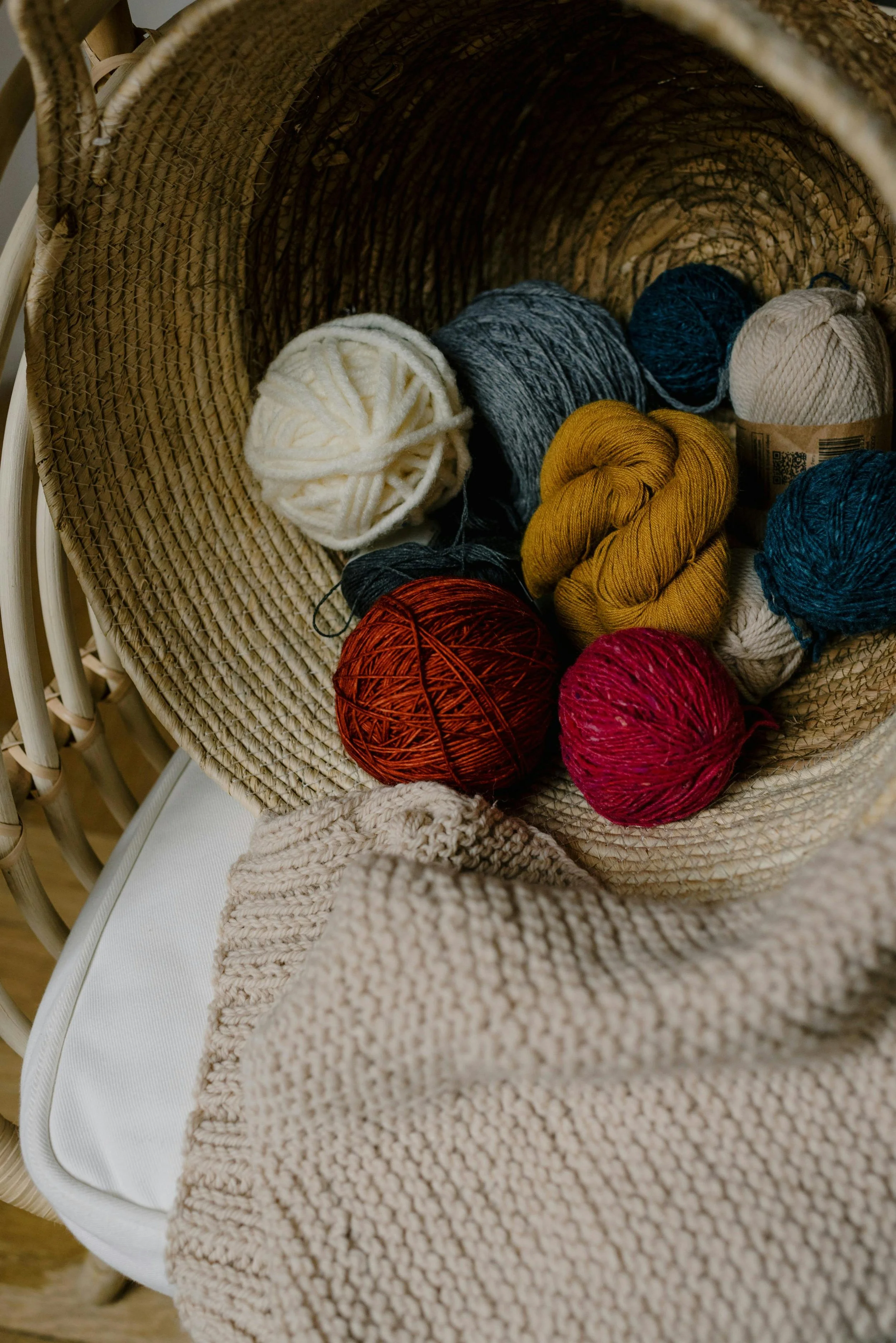 A wicker basket filled with colorful balls of yarn, including white, gray, blue, mustard yellow, red, and pink, resting on a beige knitted blanket on a white chair.