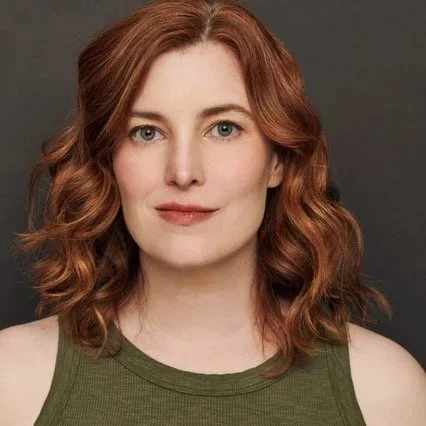 A woman with red wavy hair wearing a green sleeveless top against a dark background.