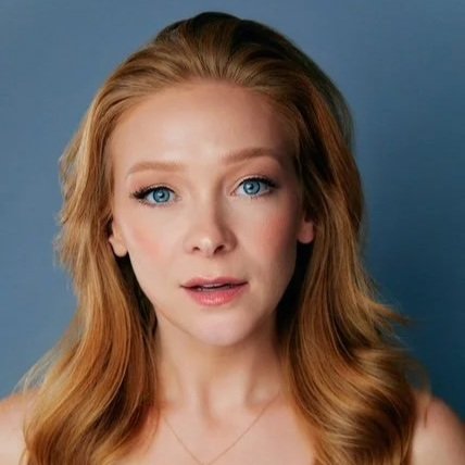 Close-up of a woman with long, wavy, red hair and blue eyes against a blue background.