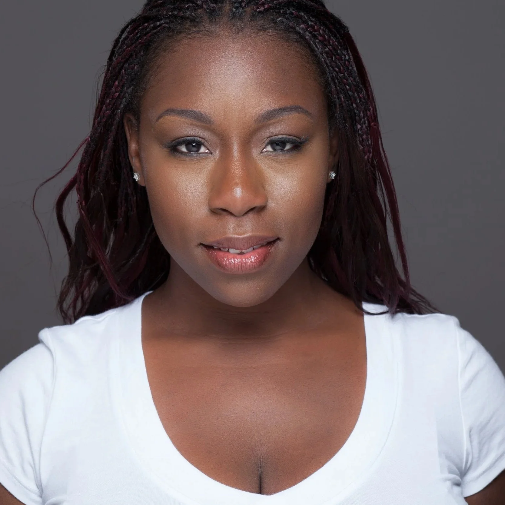 Close-up of a woman with dark skin and braided hair, wearing a white top and small pearl earrings, smiling softly against a plain gray background.
