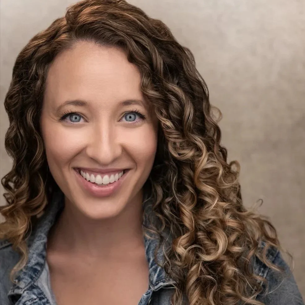 Close-up of a smiling woman with curly brown hair and blue eyes, wearing a denim jacket.