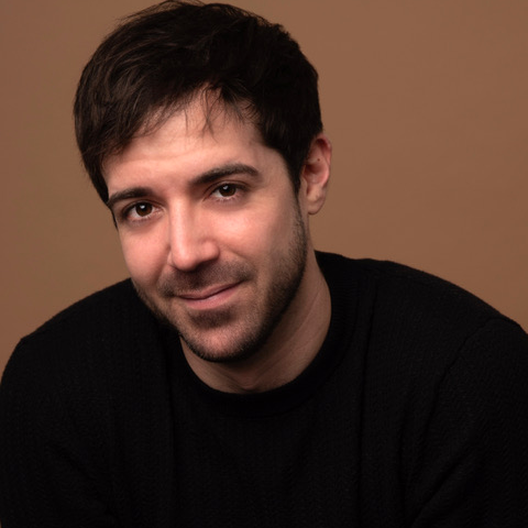 Portrait of a man with dark hair, slight beard, wearing a black sweater, against a plain brown background.