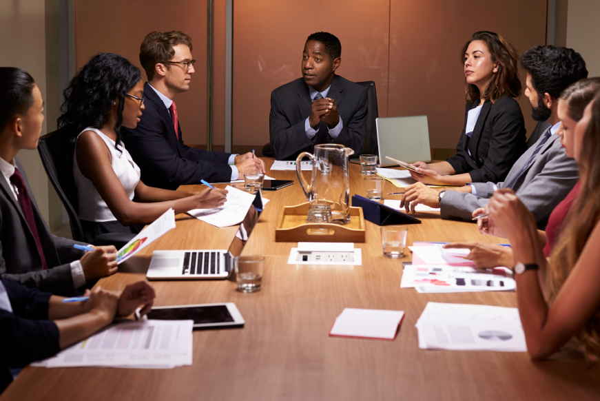 A business meeting with eight diverse professionals around a conference table, with a man speaking and others listening, some taking notes or using laptops.