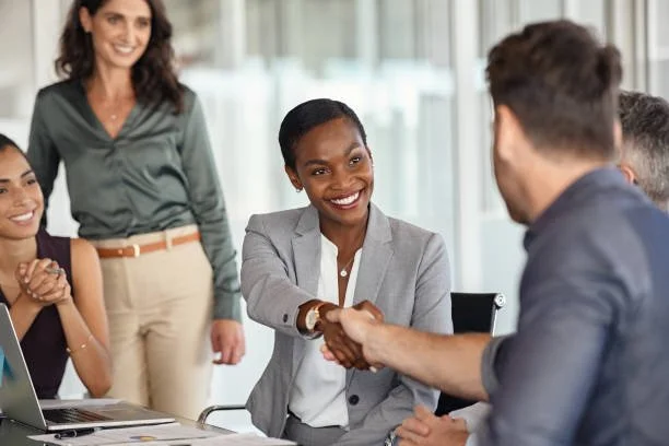 Businesswoman shaking hands with a man in a meeting room, with colleagues watching and smiling.