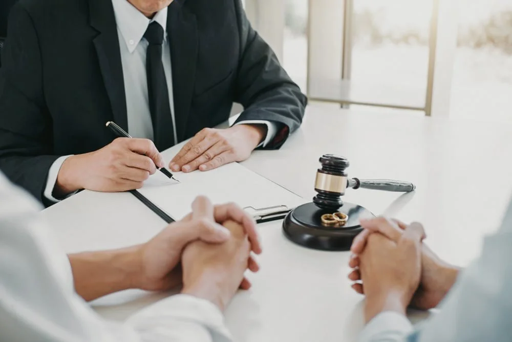 A judge in black robe and lawyers sitting at a desk during a legal proceeding, with a gavel and legal rings on the bench, in a bright office.