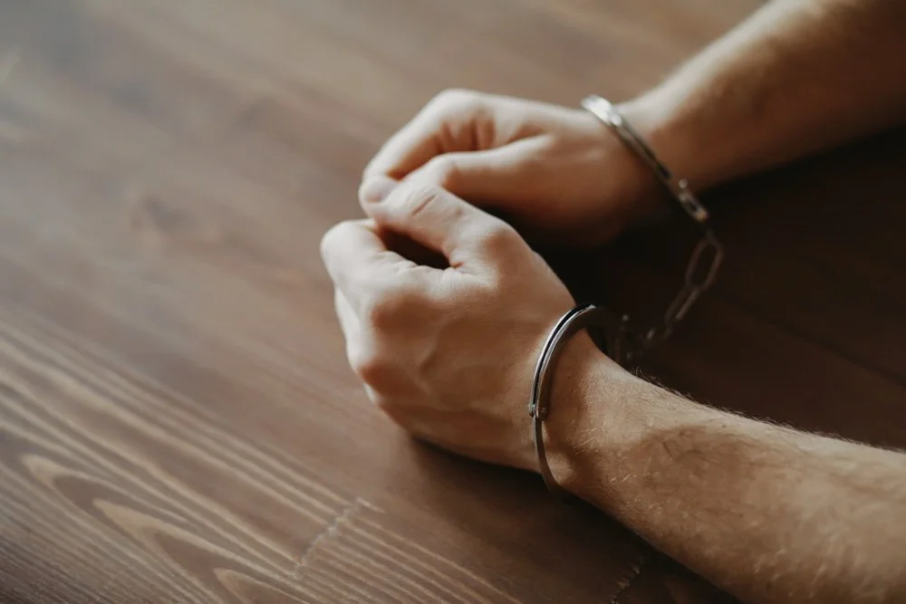 Close-up of a person's hands and wrists in handcuffs resting on a wooden surface.