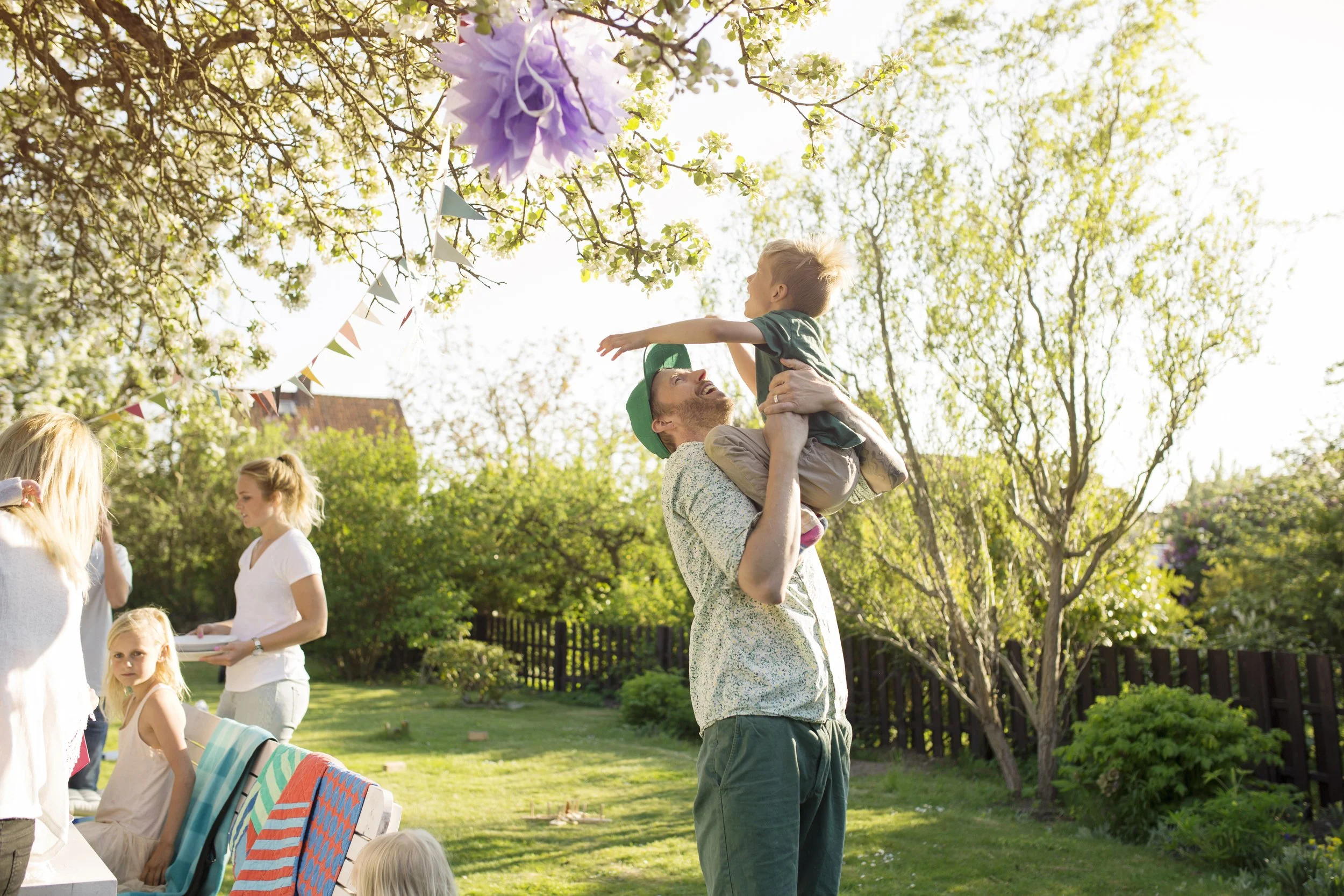 A man is lifting a young boy in the air in on a well manicured lawn. Other children and adults are nearby, some sitting and some standing, with a colorful striped towel draped over a bench.