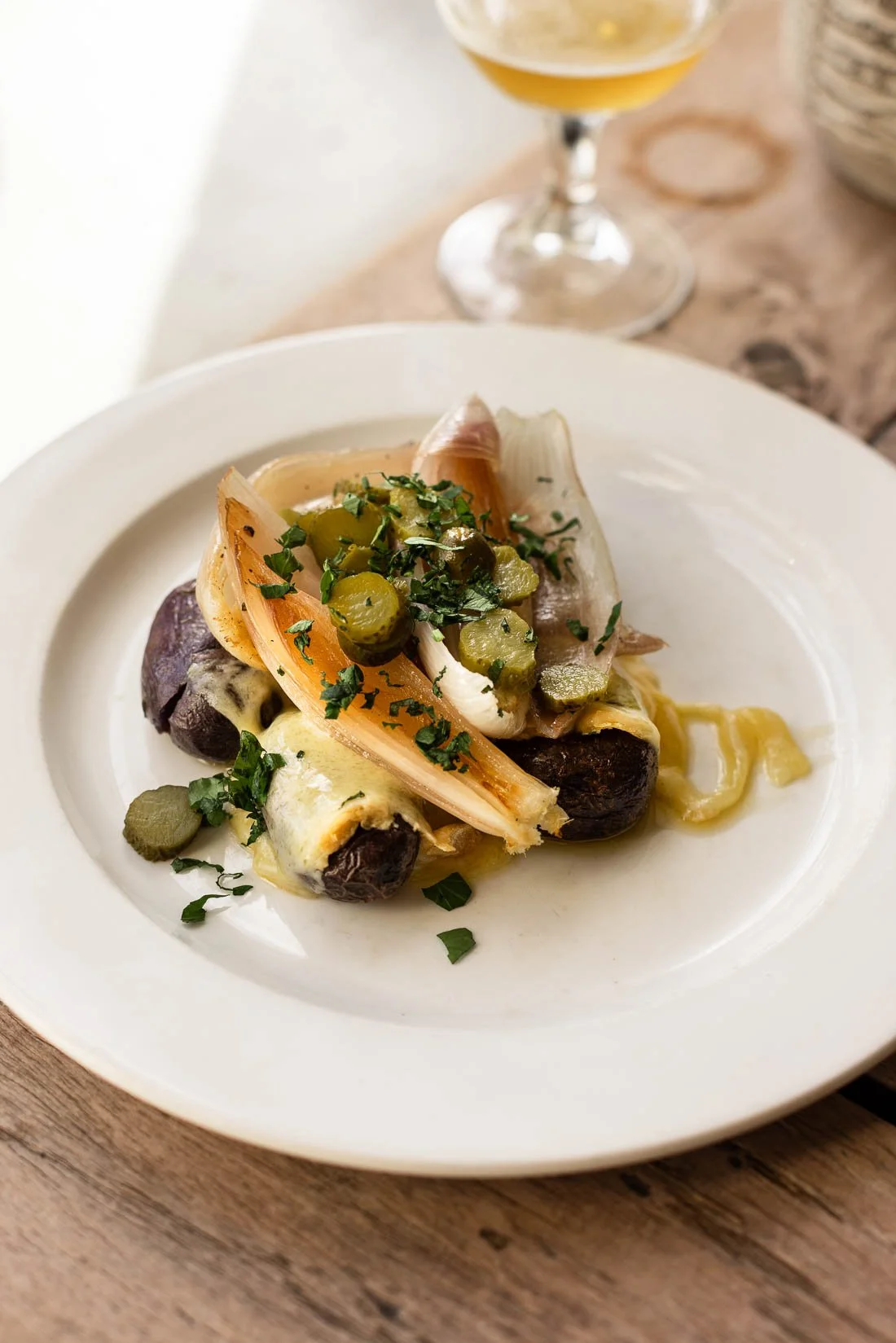 A white plate of roasted vegetables, including mushrooms, pickles, and herbs, with a glass of beer or cider in the background.