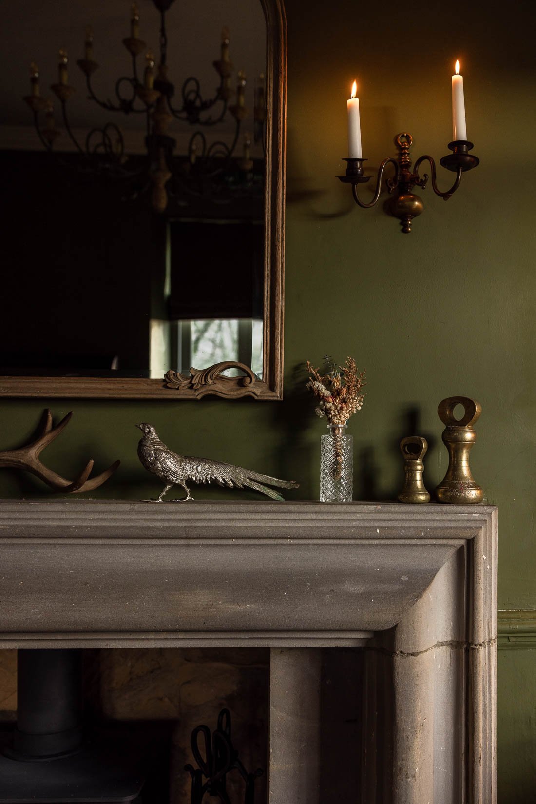 A living room decor scene featuring a fireplace mantel with decorative items, a mirror, a wall sconce with two lit candles, a small dried flower arrangement in a textured glass vase, and various metallic objects, with a reflected chandelier visible i