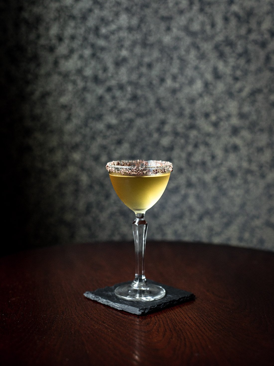 A yellow cocktail in a tall, elegant glass with a salted rim, placed on a black napkin on a wooden surface, against a gray textured background.