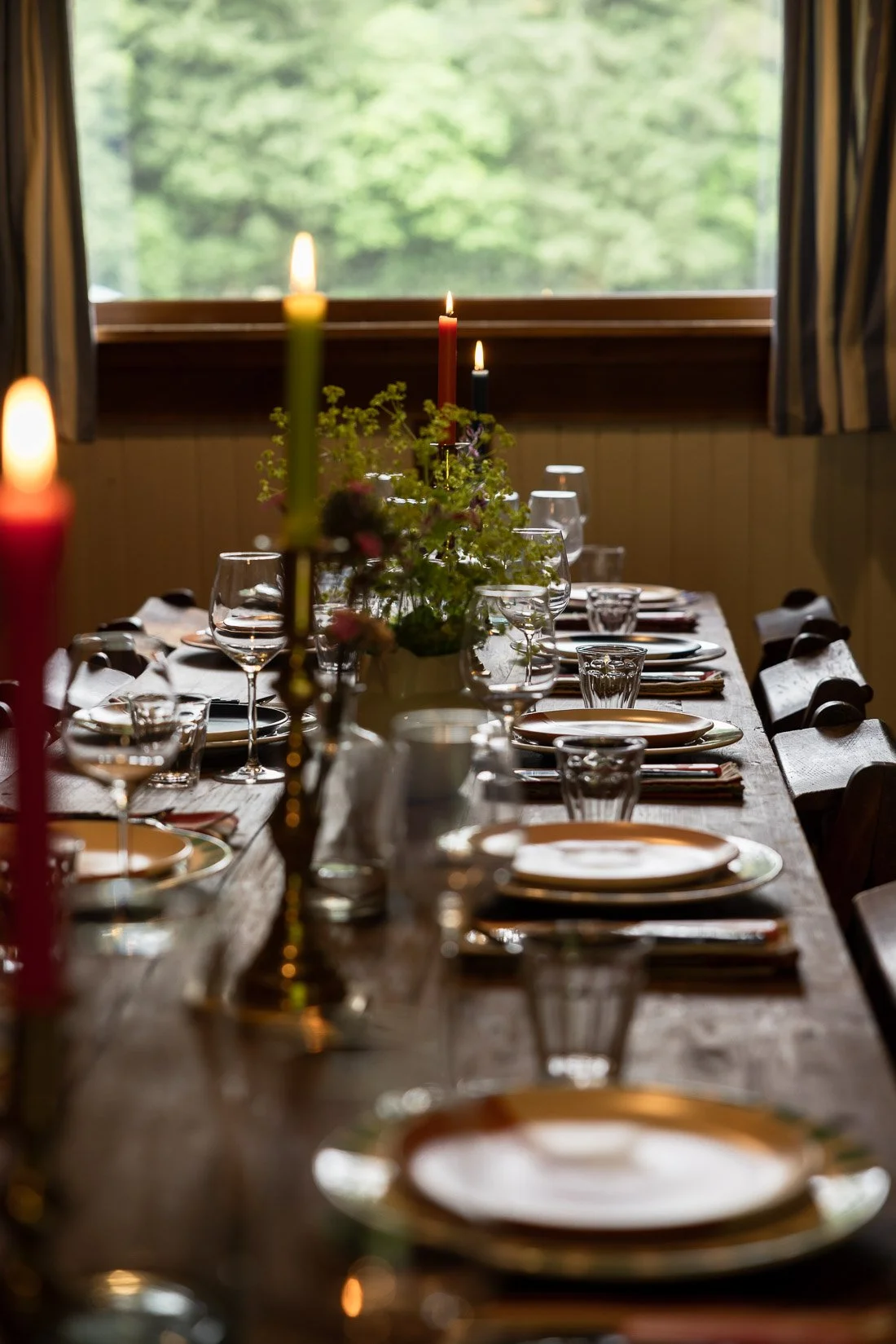 A dining table set for a meal with plates, glasses, and silverware, featuring a centerpiece with flowers and lit candles, in front of a window with a green outdoor view.