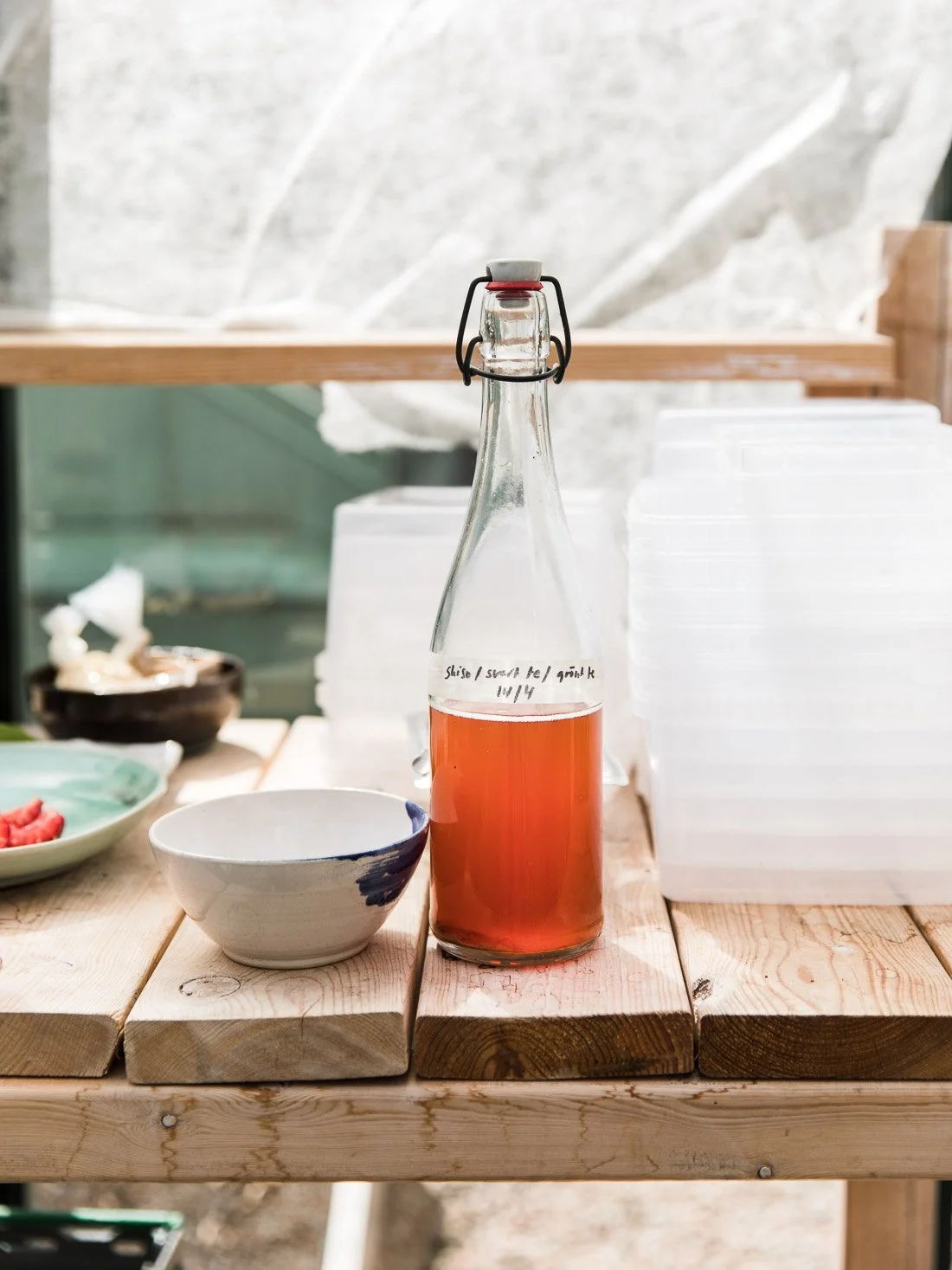 A glass bottle filled with pinkish-orange liquid on a wooden table, with a white bowl nearby and stacked white containers in the background.