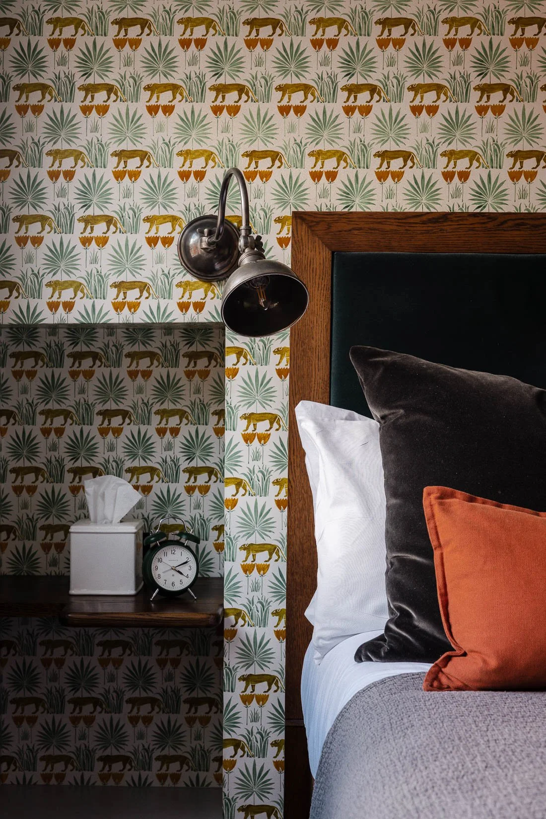 A bedroom scene featuring a wooden headboard with black and orange pillows, a wall with tropical animal and plant wallpaper, a black wall-mounted lamp, a small bedside table with a tissue box and an alarm clock.