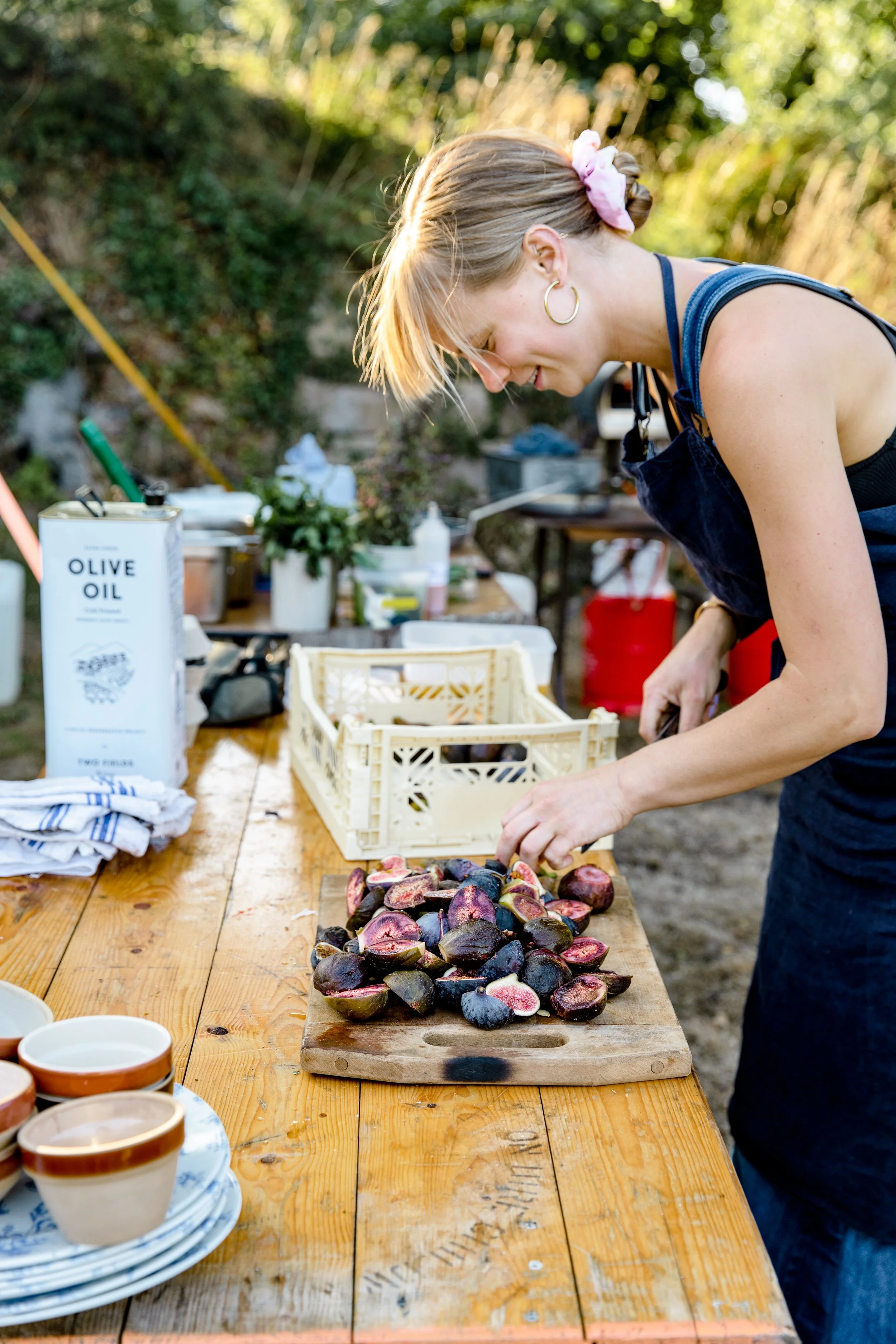 A woman preparing fresh figs on a wooden outdoor table, with a variety of dishes and utensils nearby in the background, during daylight.