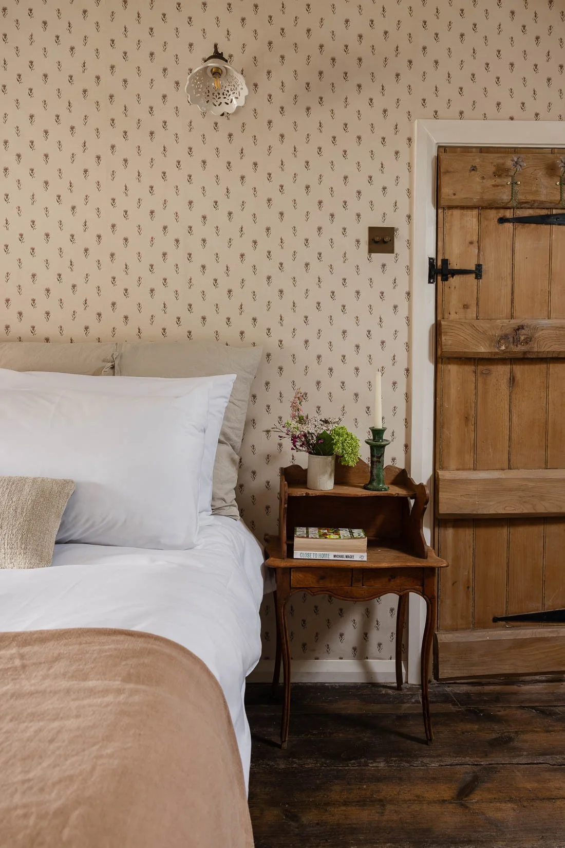 A bedroom corner with a bed, a vintage wooden nightstand with flowers, books, and a candle holder, a wooden door, and wallpaper with a small floral pattern.