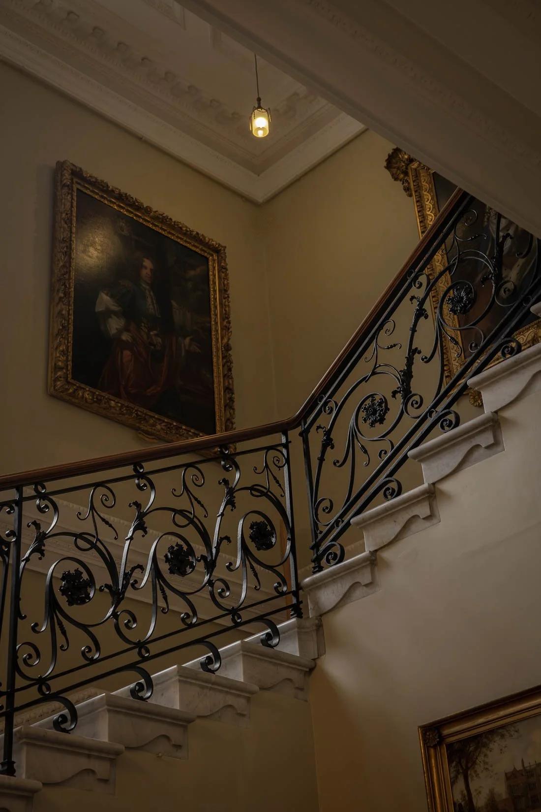 Interior view of a staircase with ornate black wrought iron railing, a large framed portrait on the wall, and a ceiling light fixture inside a classical building.